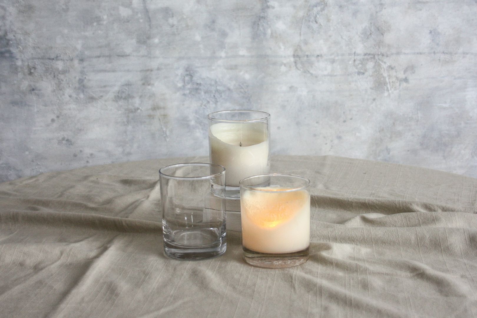 Image of three white RURAL candles in clear glass containers at various stages of burning, sitting on top of an earthy-toned linen cloth and set agains a grey painted canvas. The vibe is very cool artist studio.