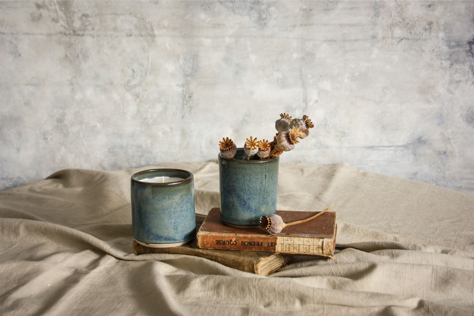 Photo is of two beautifully handmade pottery candle cups in a blue green and brown glaze, sitting on top of vintage books and an earthy toned linen cloth. There are dried poppy blossoms in one cup, and a grey abstract painting creates the backdrop. The vibe is very much a vintage artists studio.