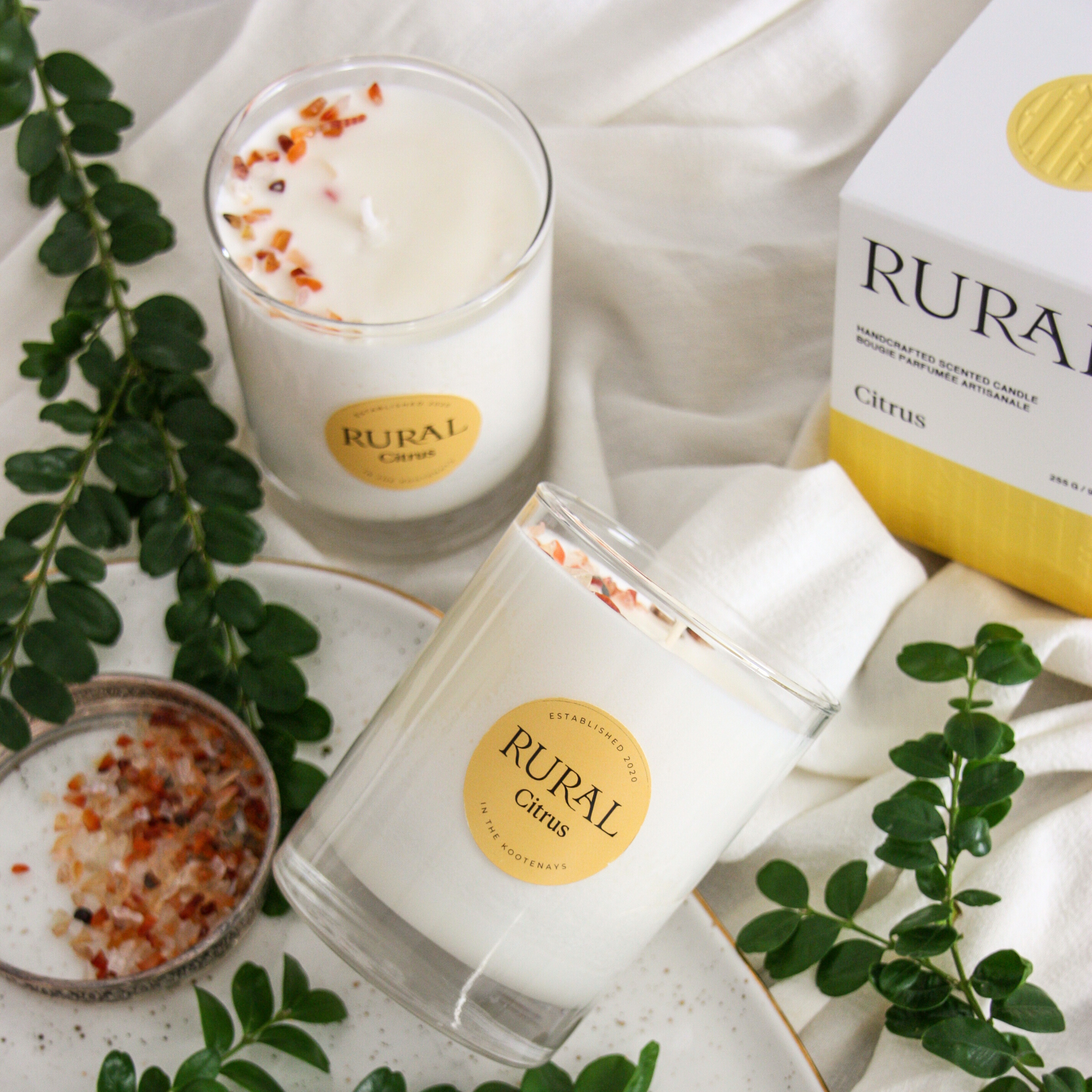 Photo of two white candles in clear glass containers with yellow round RURAL Candles labels on, indicating the scent is called Citrus. We can the top of the candle is embellished with orange carnelian gemstones, beautifully arranged in a half-moon pattern. We can also see the matching white and yellow RURAL Candles gift box for the Citrus Candle.