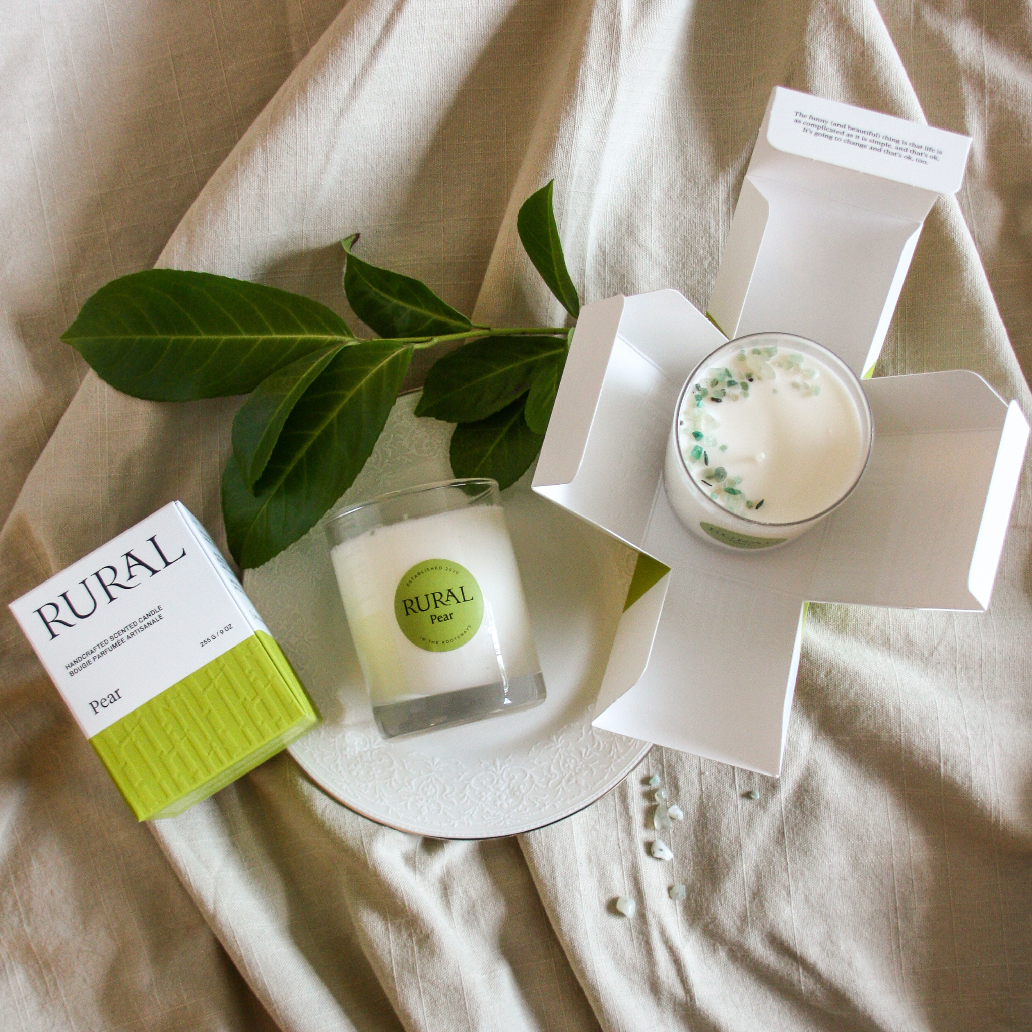 Top view photo of a white candle in a clear glass with a round green label indicating RURAL Candles Pear Scent beside its matching green and white elegantly embossed candle gift box. We can see the top of the candle is decorated with small green gemstones, artfully arranged in a crescent moon shape. The gift box opens up like a flower.