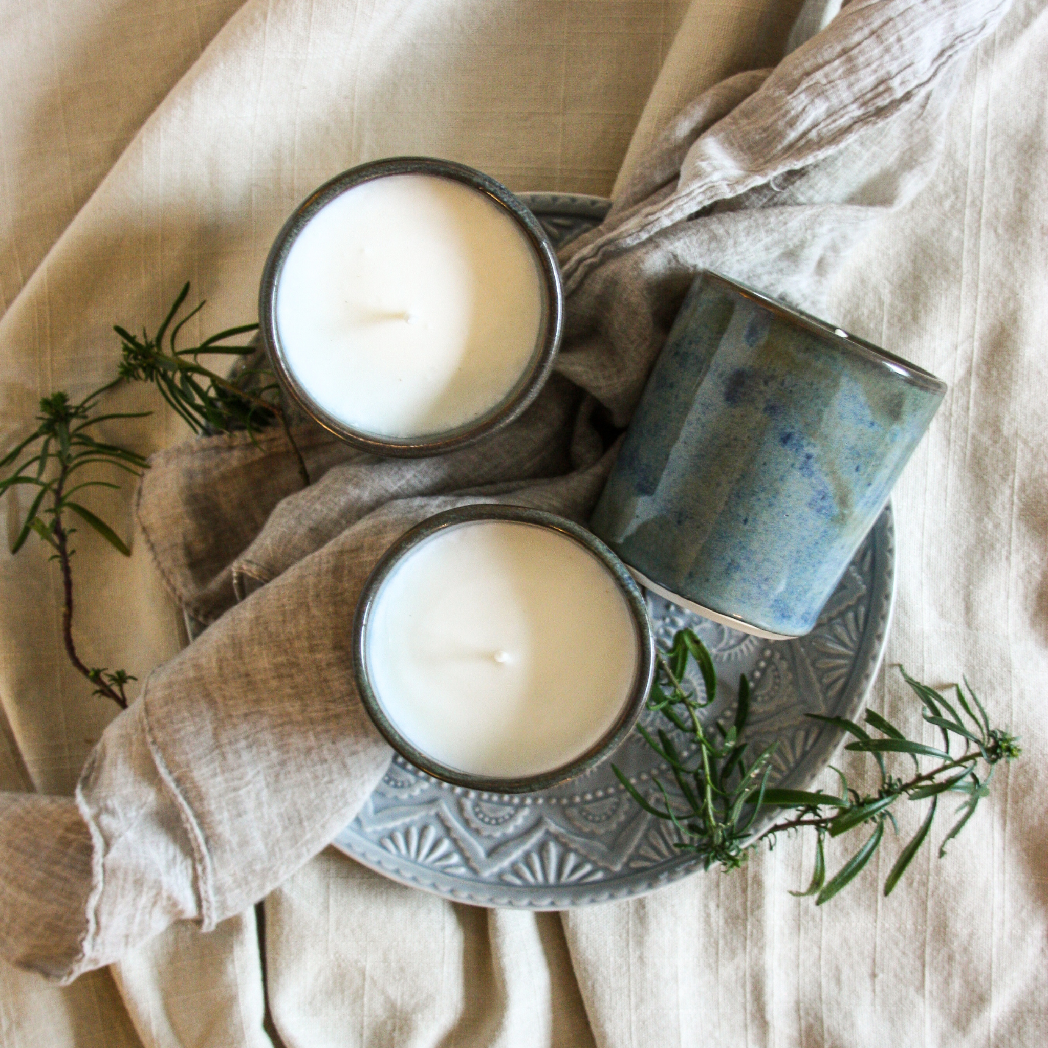 Photo of a group of three handcrafted pottery candles with a beautiful blue-green and brown glaze, arranged on rustic grey ceramic plate with earth-toned linens and green herbs surrounding them. The vibe is very artistic, serene and inviting kitchen space.