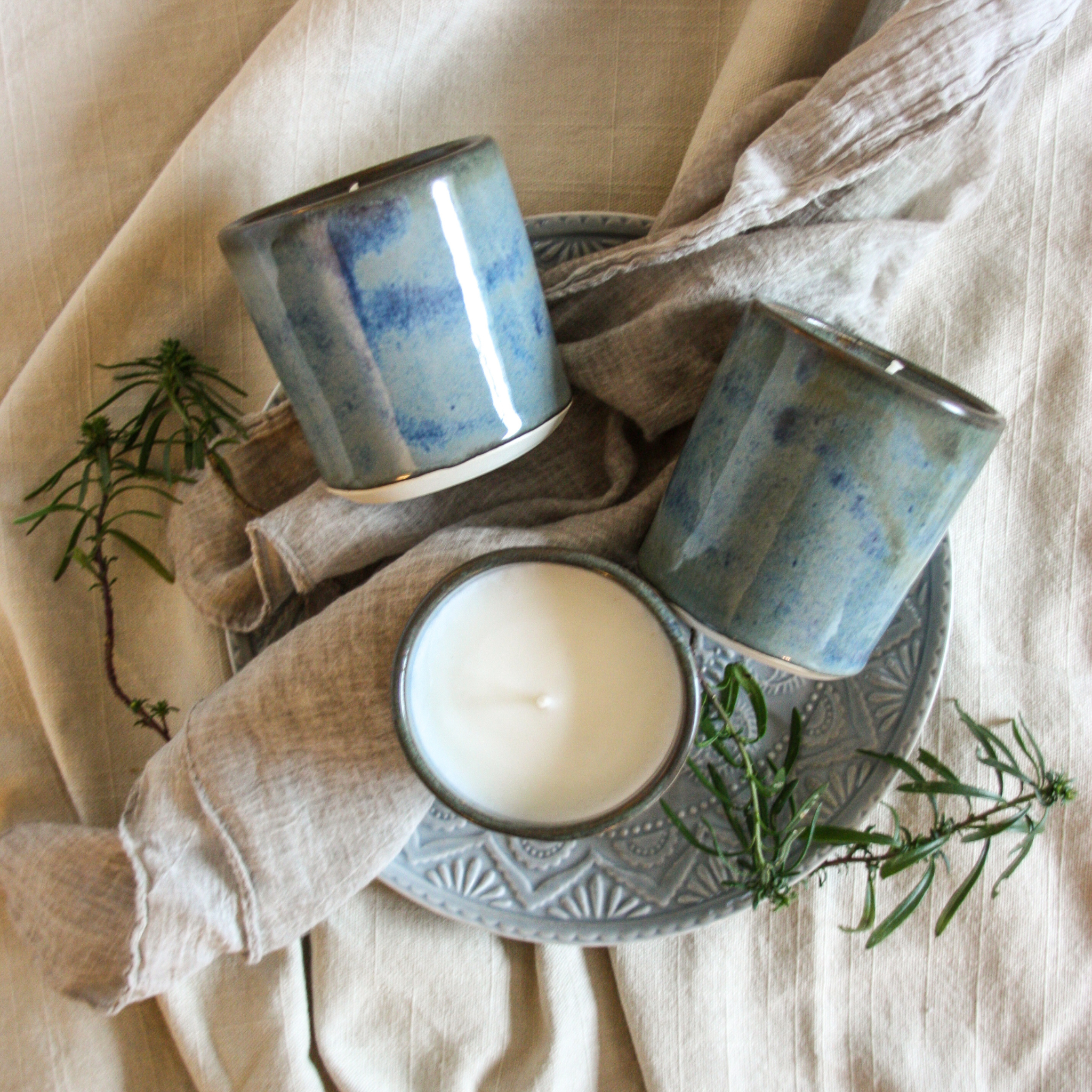 Photo of a group of three handcrafted pottery candles with a beautiful blue-green and brown glaze, arranged on rustic grey ceramic plate with earth-toned linens and green herbs surrounding them. The vibe is very artistic, serene and inviting kitchen space.