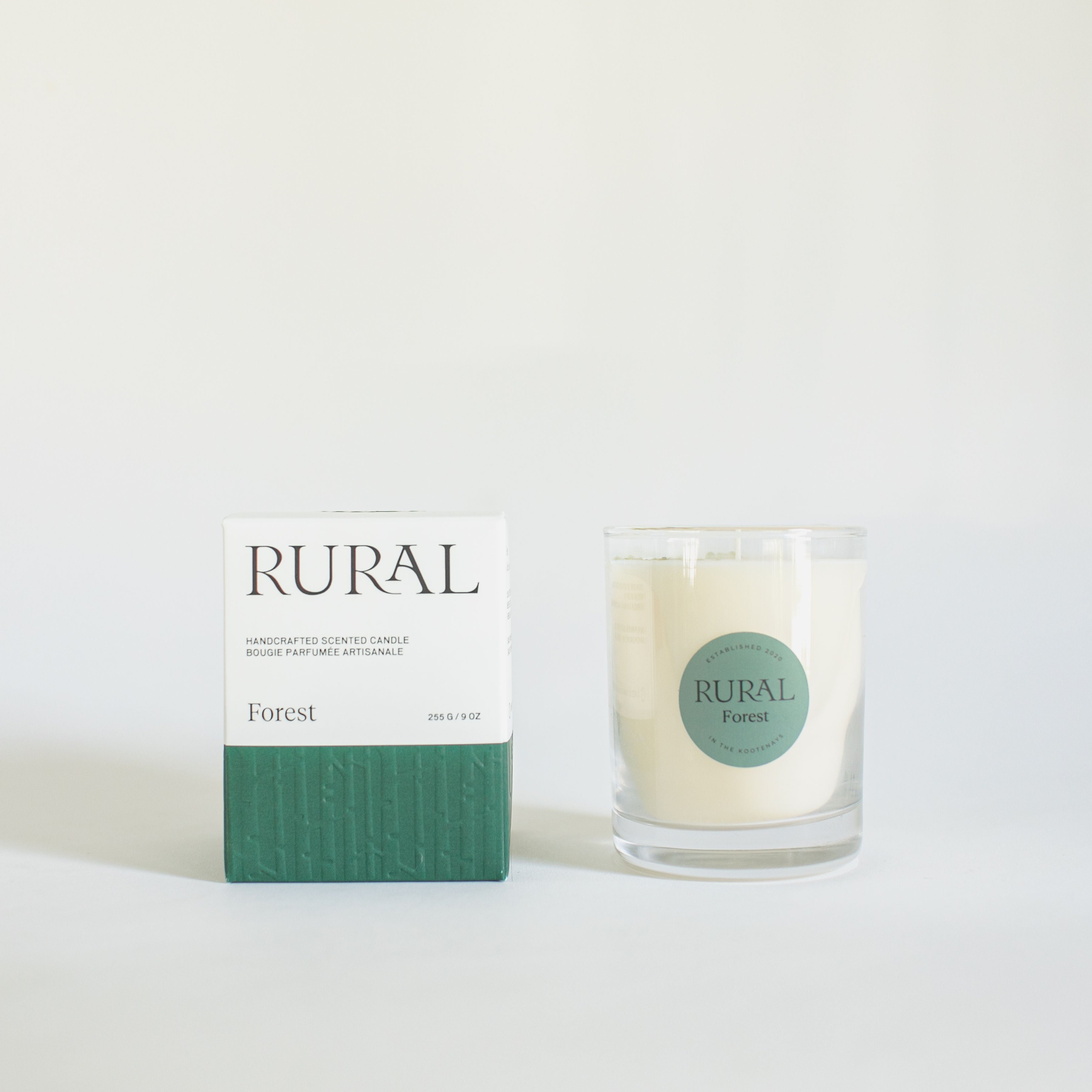 Photo of a white candle in a clear glass with a round green label indicating RURAL Candles Forest Scent beside its matching green and white elegantly embossed candle gift box.