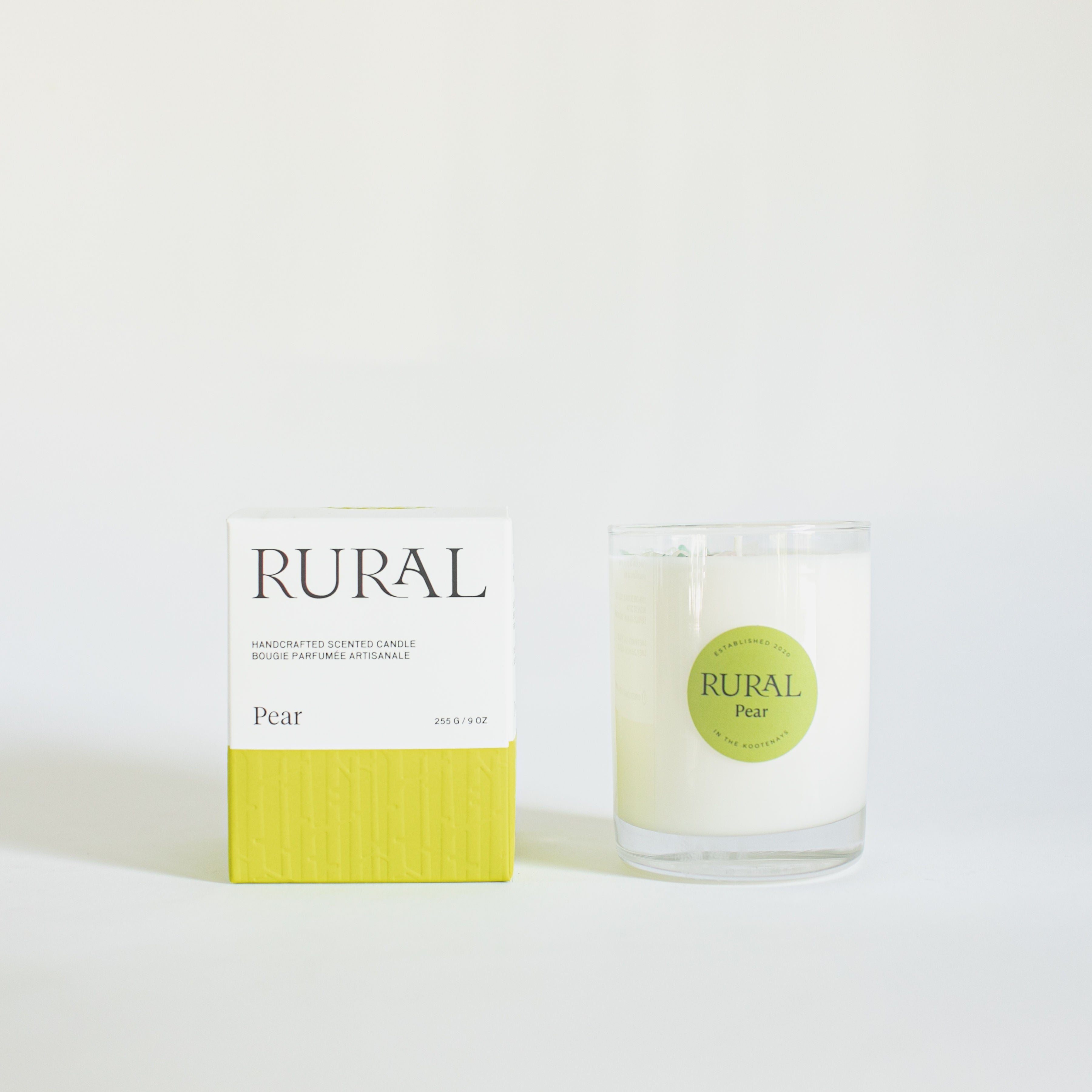 Photo of a white candle in a clear glass with a round green label indicating RURAL Candles Pear Scent beside its matching green and white elegantly embossed candle gift box.