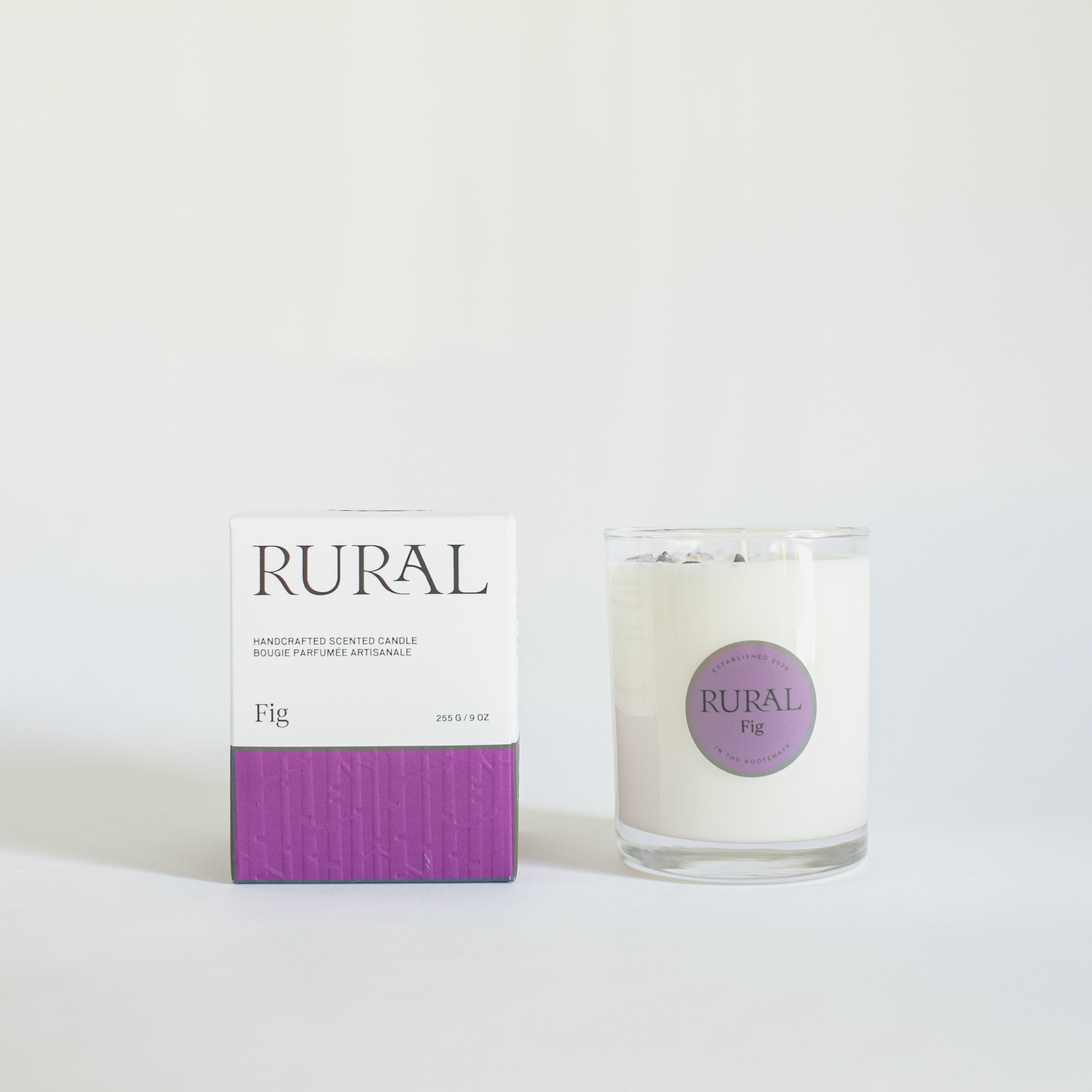 Photo of a white candle in a clear glass with a round purple label indicating RURAL Candles Fig Scent beside its matching purple and white elegantly embossed candle gift box.