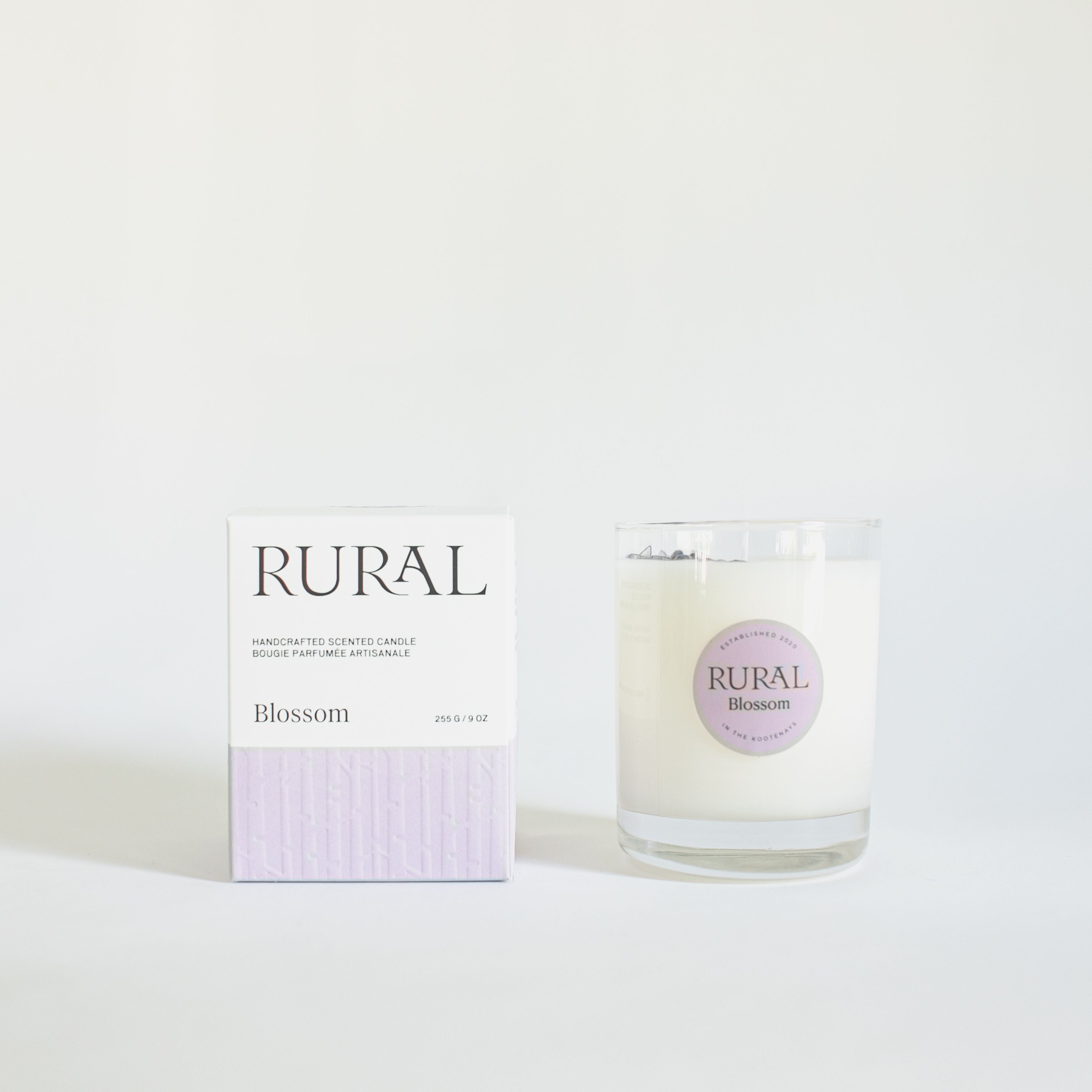 Photo of a white candle in a clear glass container with a light purple round RURAL Candles label on it, indicating the scent is called Blossom. The candle top is embellished with a sprinkling of purple amethyst chips, and the candle is sitting beside its matching purple and white RURAL Candles gift box.