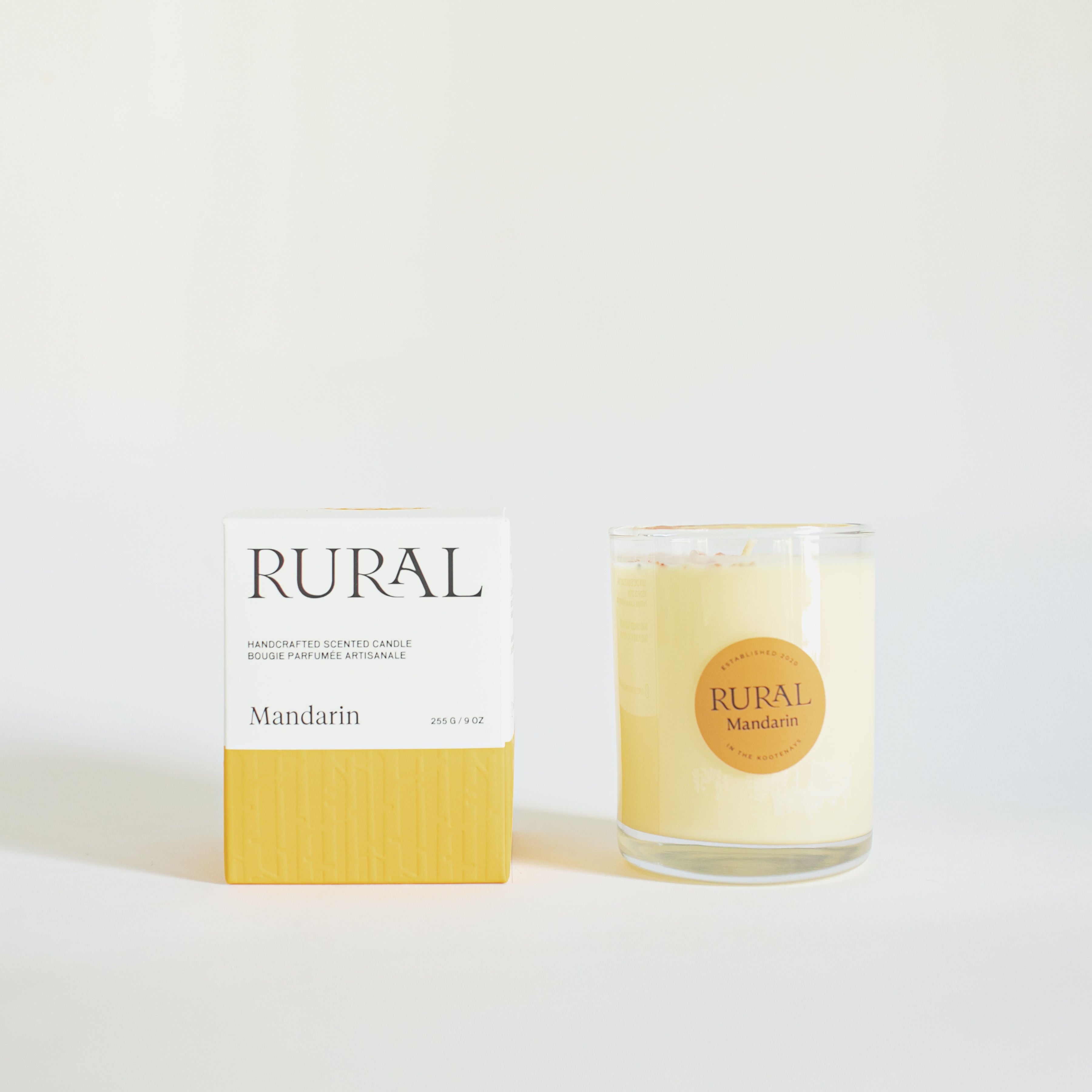 Photo of a coconut soy handcrafted scented candle in a clear glass with a round orange label indicating RURAL Candles Mandarin Scent beside its matching orange and white embossed candle gift box.