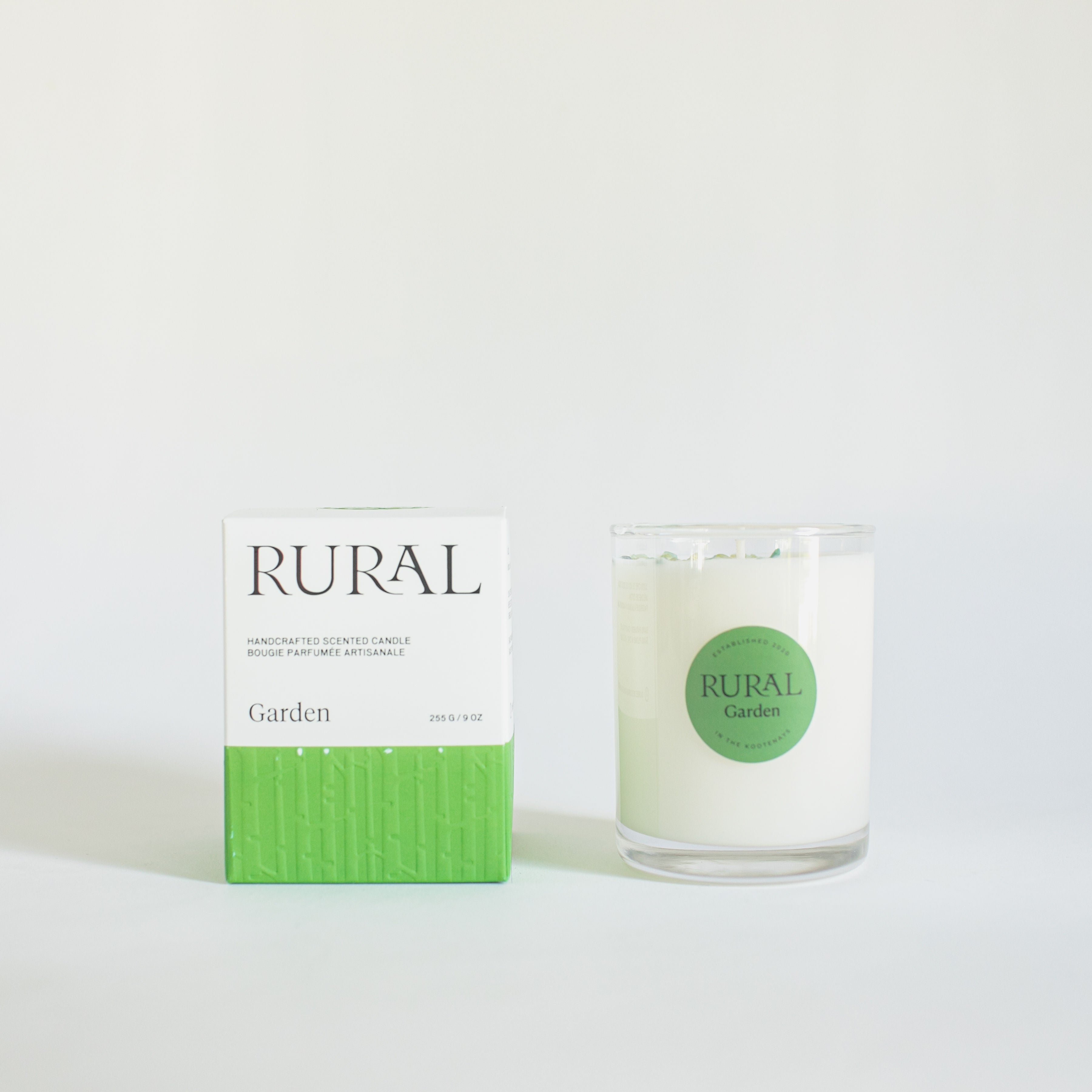 Photo of a white candle in a clear glass with a round green label indicating RURAL Candles Garden Scent beside its matching green and white elegantly embossed gift box.