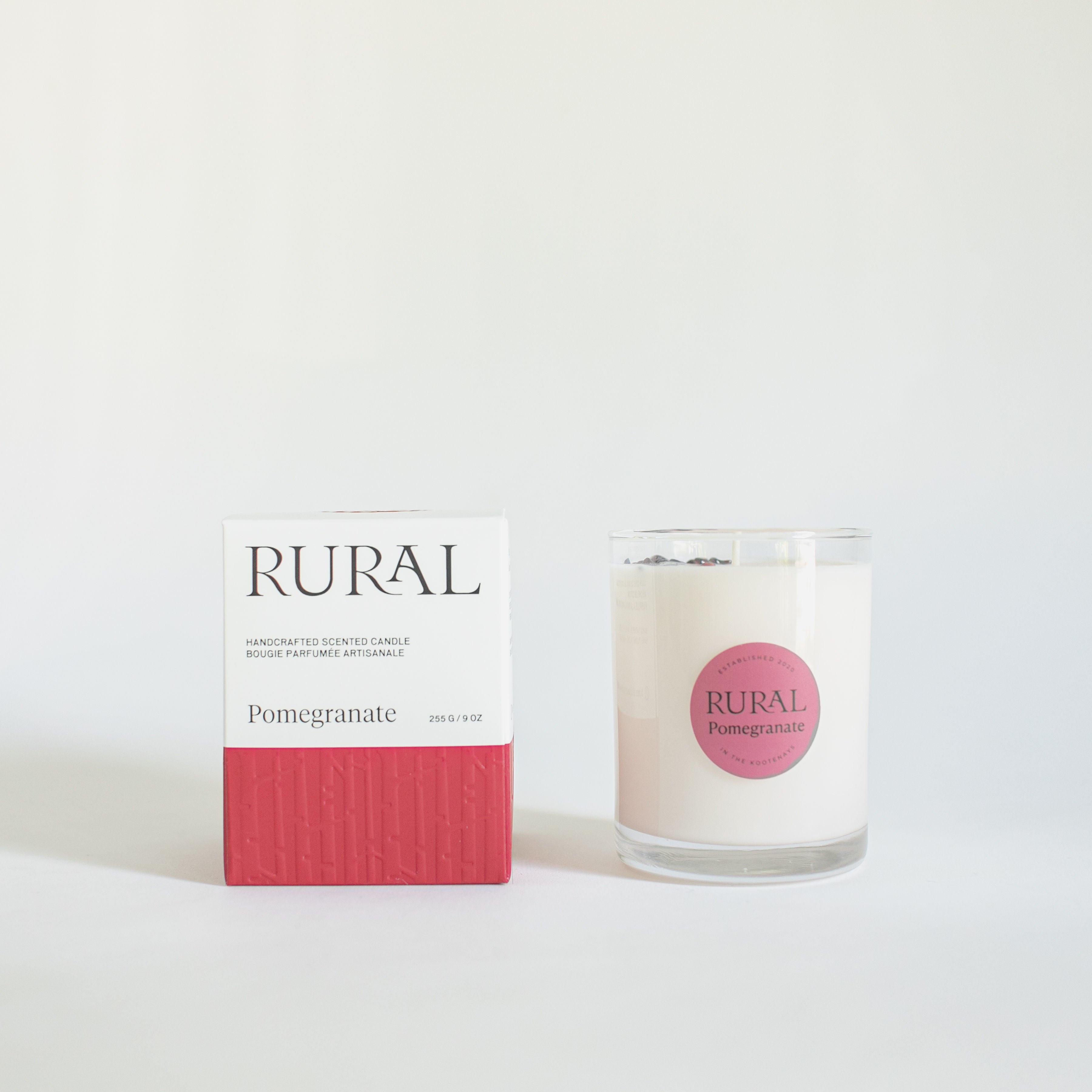 Photo of a white candle in a clear glass with a round red label indicating RURAL Candles Pomegranate Scent beside its matching red and white elegantly embossed candle gift box.