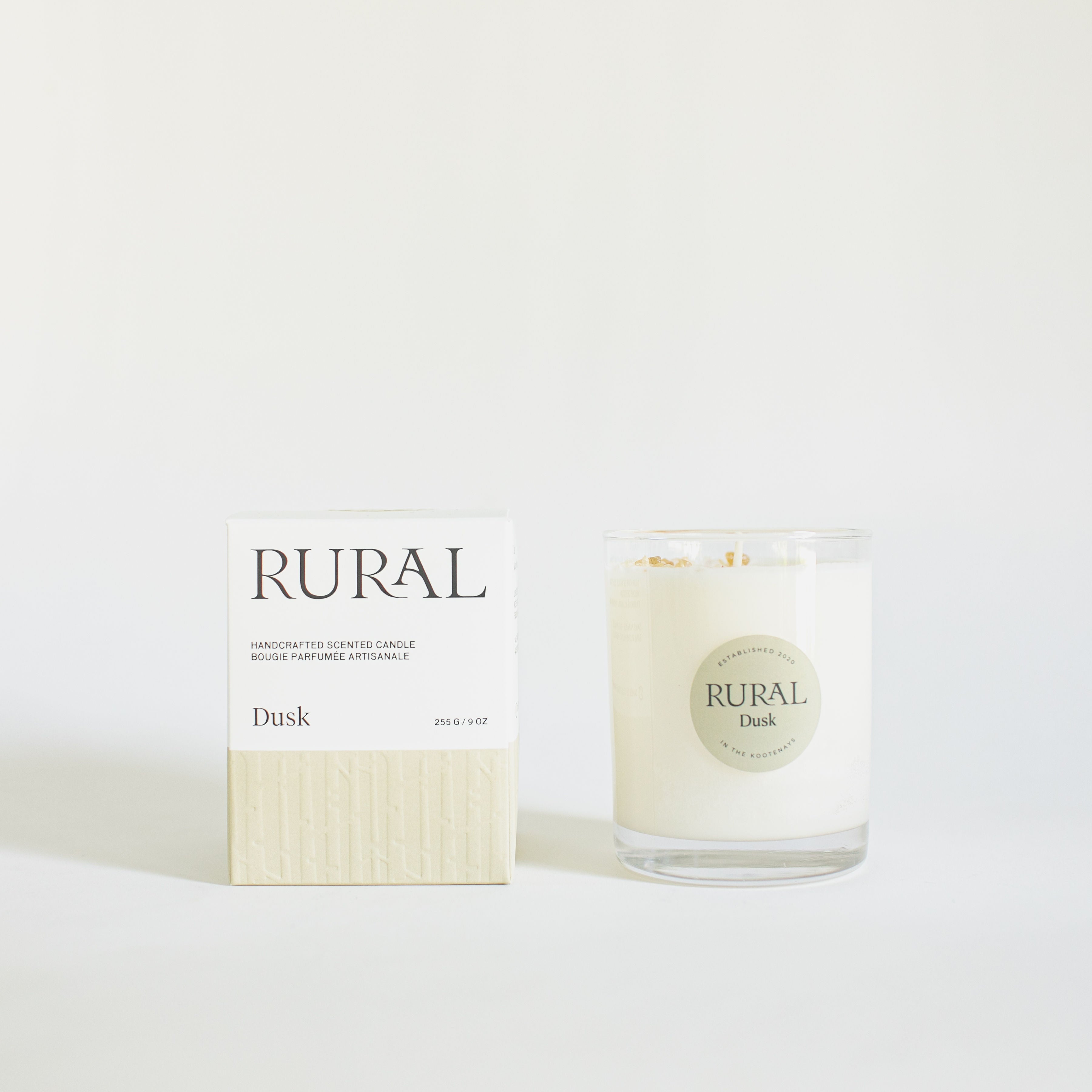 Photo of a white candle in a clear glass with a round tan label indicating RURAL Candles Dusk Scent beside its matching tan and white elegantly embossed candle gift box.