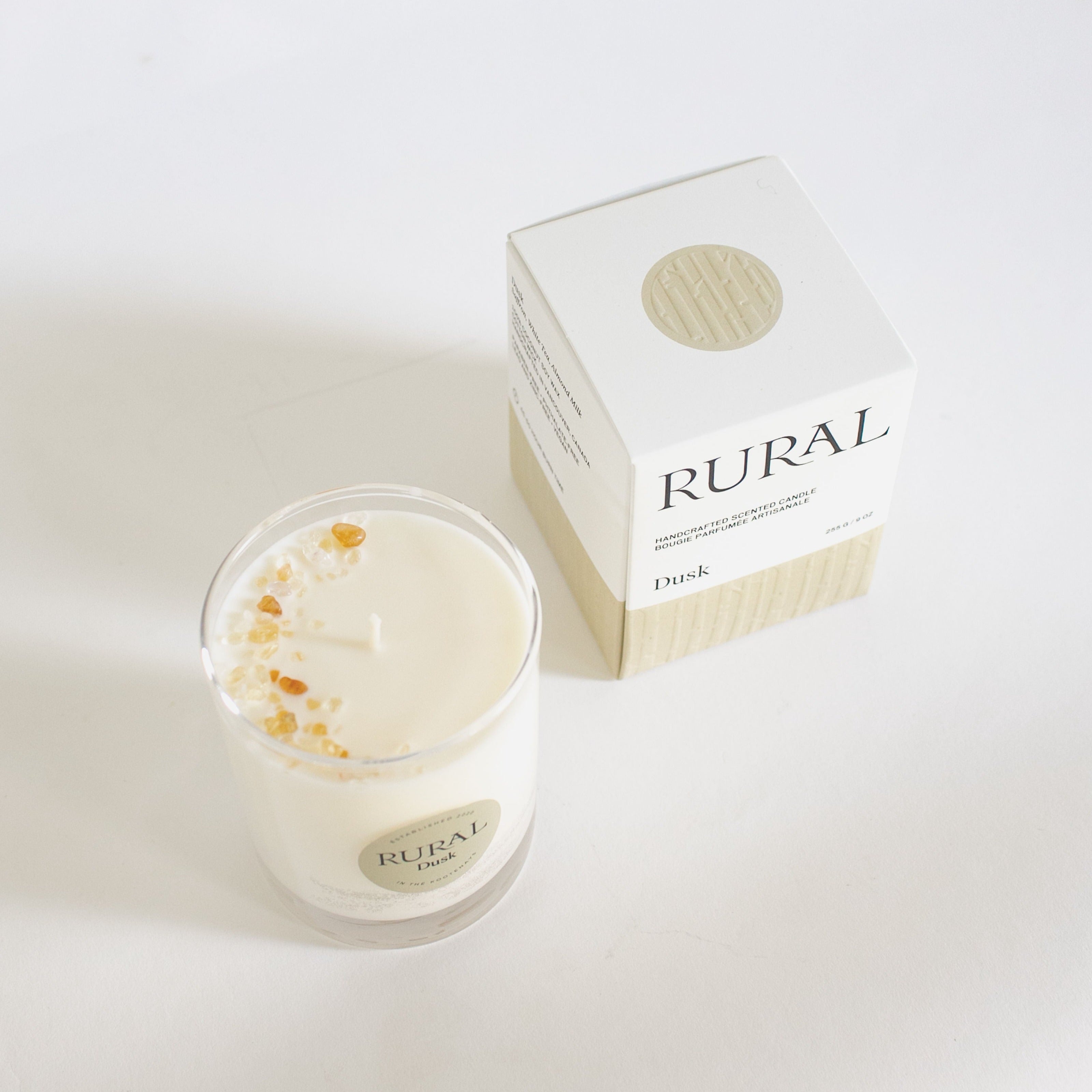 Top view photo of a white candle in a clear glass with a round tan label indicating RURAL Candles Dusk Scent beside its matching tan and white elegantly embossed candle gift box. We can see the top of the candle is decorated with small gold gemstones, artfully arranged in a crescent moon shape.