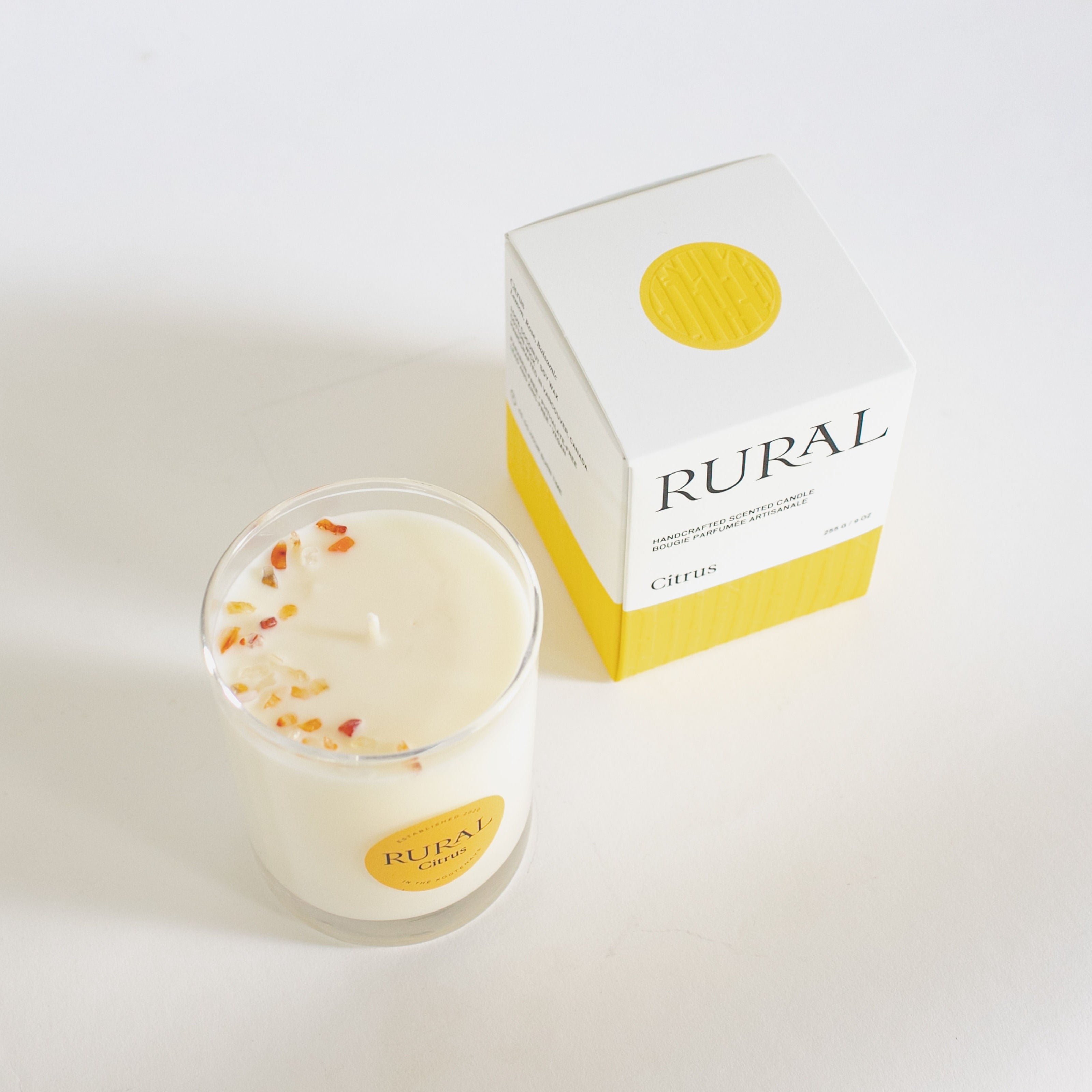 Top view of the product photo of a Citrus candle in a clear glass container with a yellow round RURAL Candles label on it, alongside its matching yellow and white RURAL Candles gift box. We can see the small orange carnelian gemstones, all artfully arranged on the top of the candle.