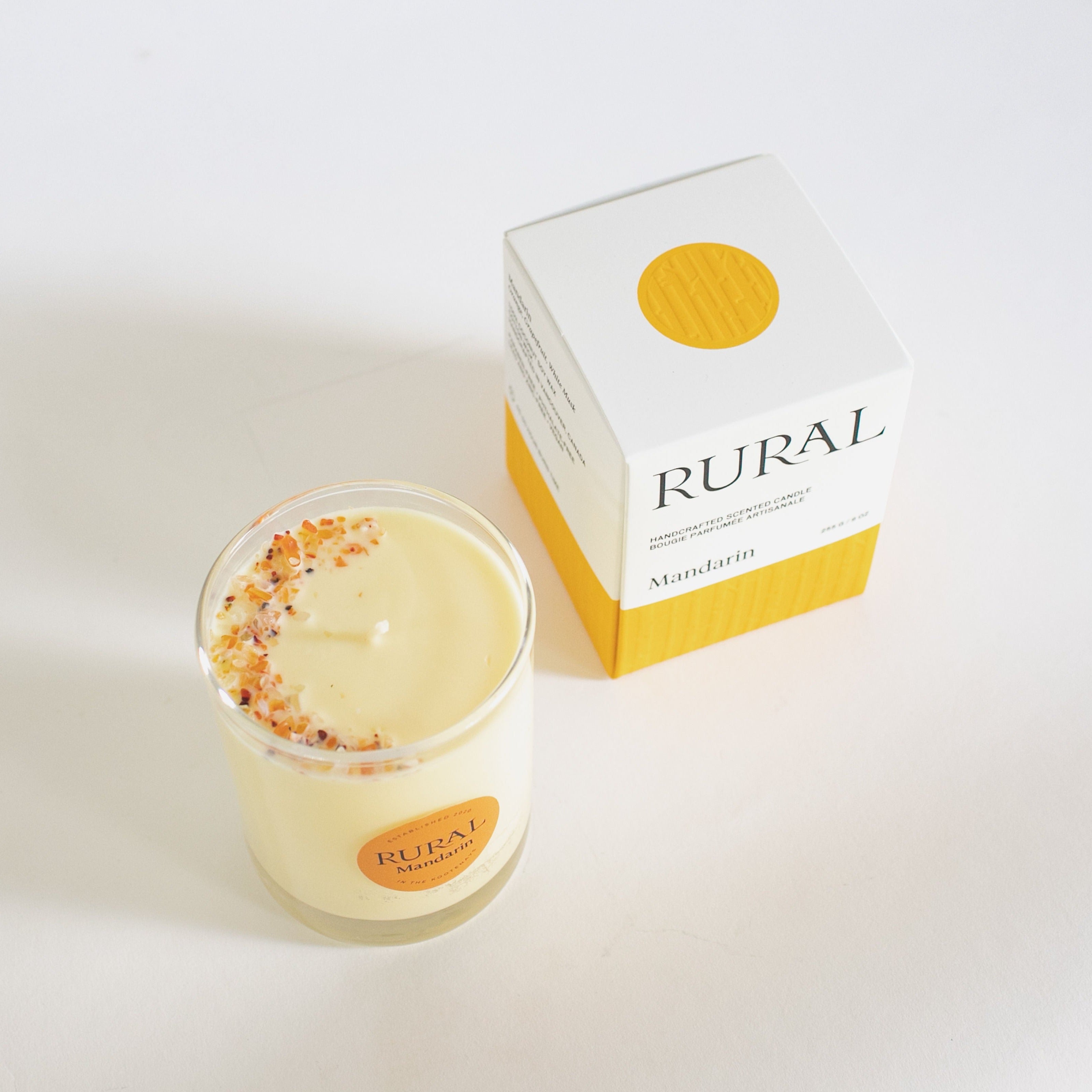 Top view photo of a candle in a clear glass with a round orange label indicating RURAL Candles Mandarin Scent beside its matching orange and white embossed candle gift box. We can see the top of the candle is decorated with small orange carnelian gemstones, artfully arranged in a crescent moon shape.