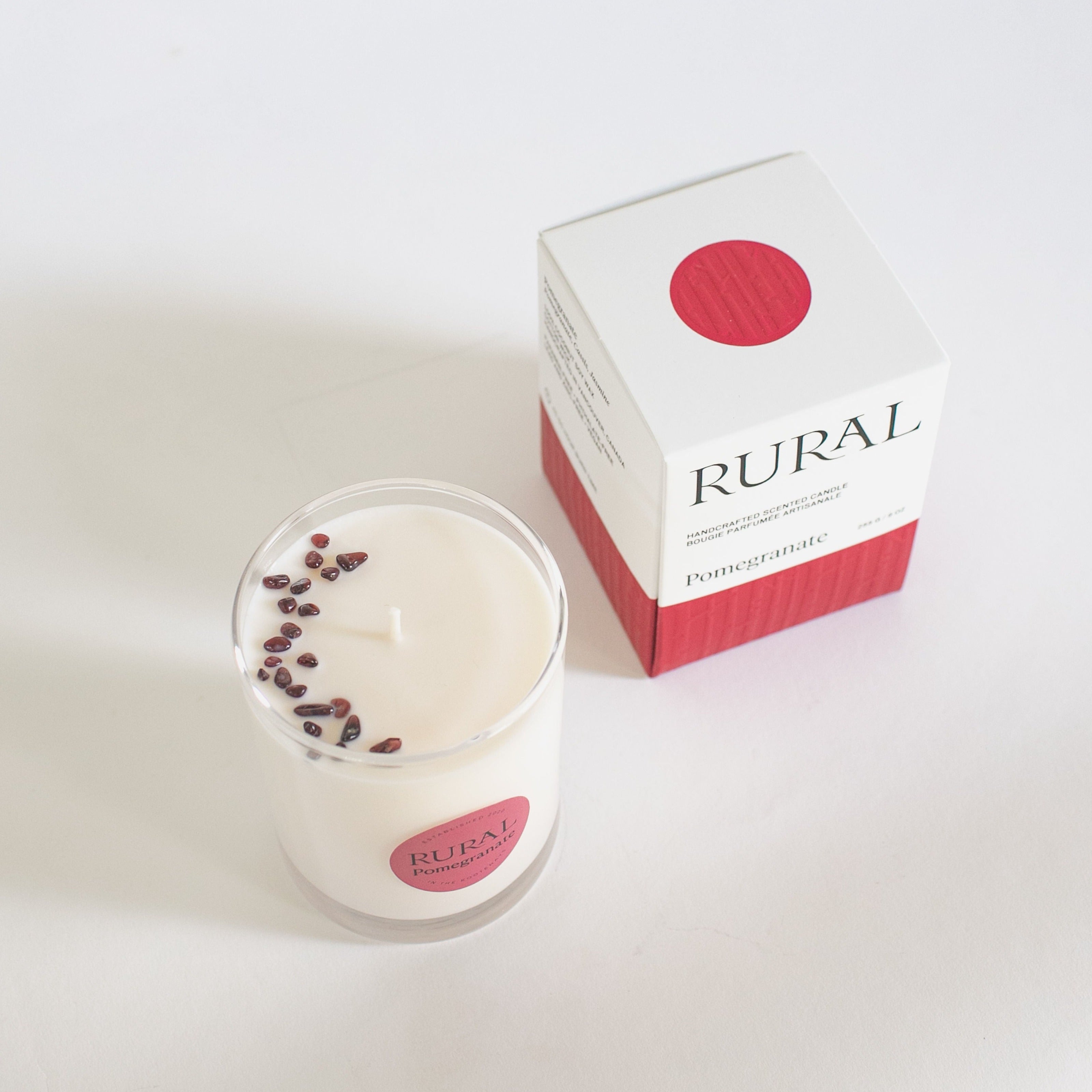Top view photo of a white candle in a clear glass with a round red label indicating RURAL Candles Pomegranate Scent beside its matching red and white elegantly embossed candle gift box. We can see the top of the candle is decorated with small red garnet gemstones, artfully arranged in a crescent moon shape.