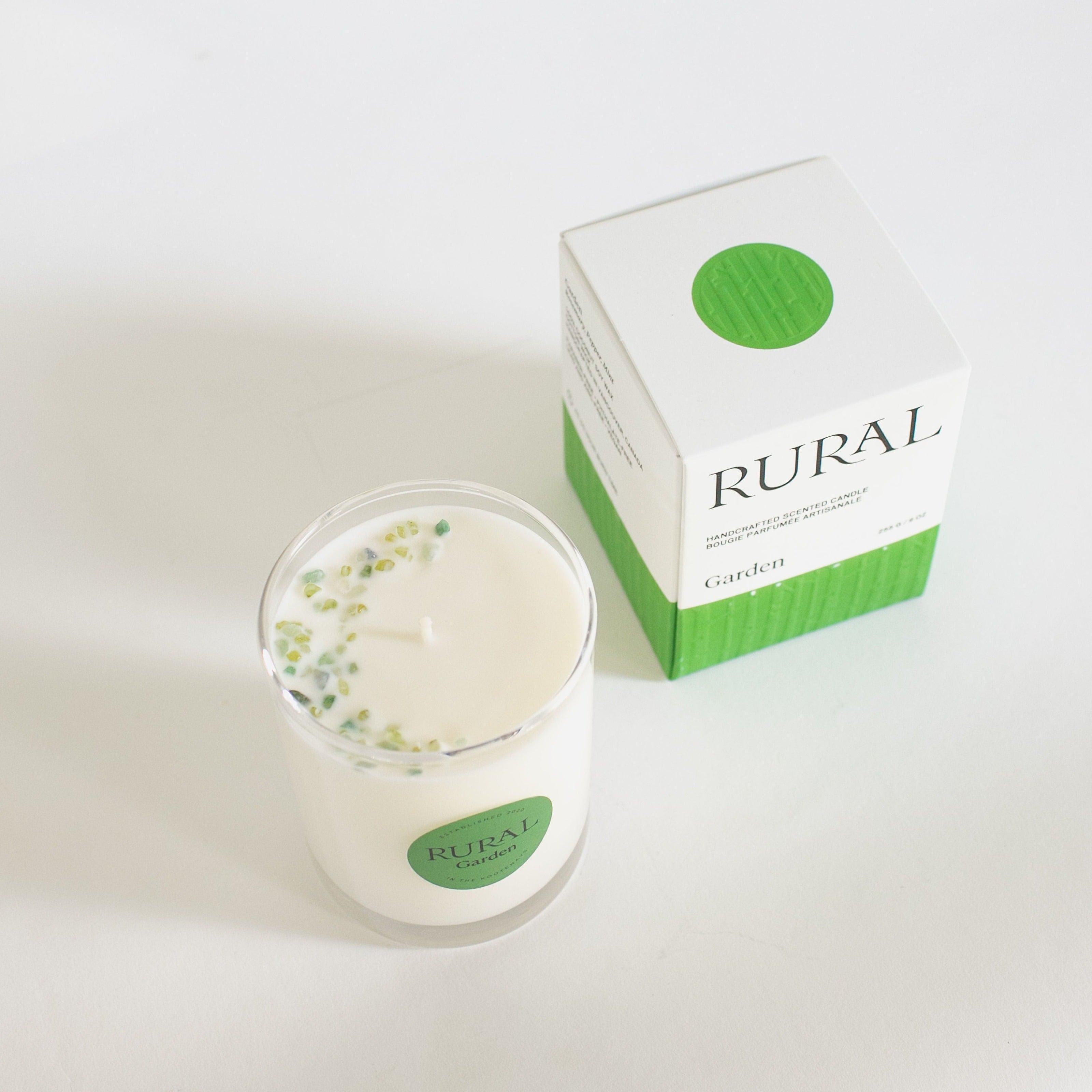 Top view photo of a white candle in a clear glass with a round green label indicating RURAL Candles Garden Scent beside its matching green and white elegantly embossed gift box. We can see the top of the candle is decorated with small green gemstones, artfully arranged in a crescent moon shape.