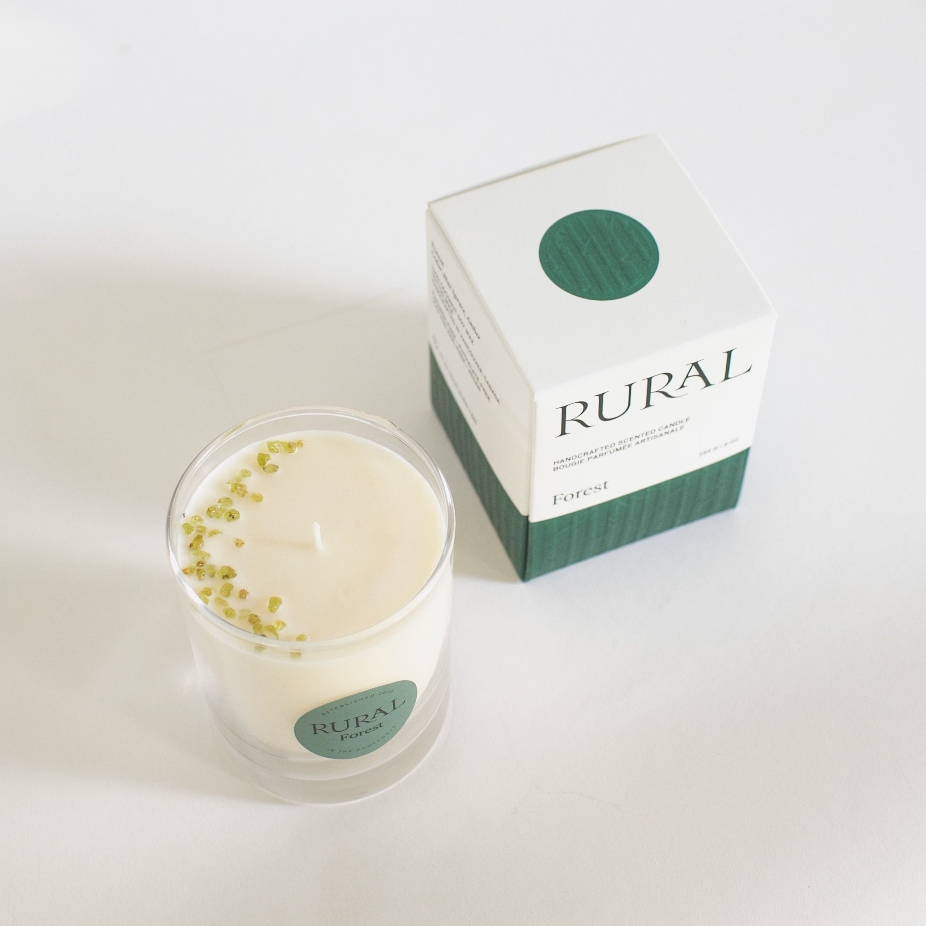 Top view photo of a white candle in a clear glass with a round green label indicating RURAL Candles Forest Scent beside its matching green and white elegantly embossed candle gift box. We can see the top of the candle is decorated with small green olivine gemstones, artfully arranged in a crescent moon shape.