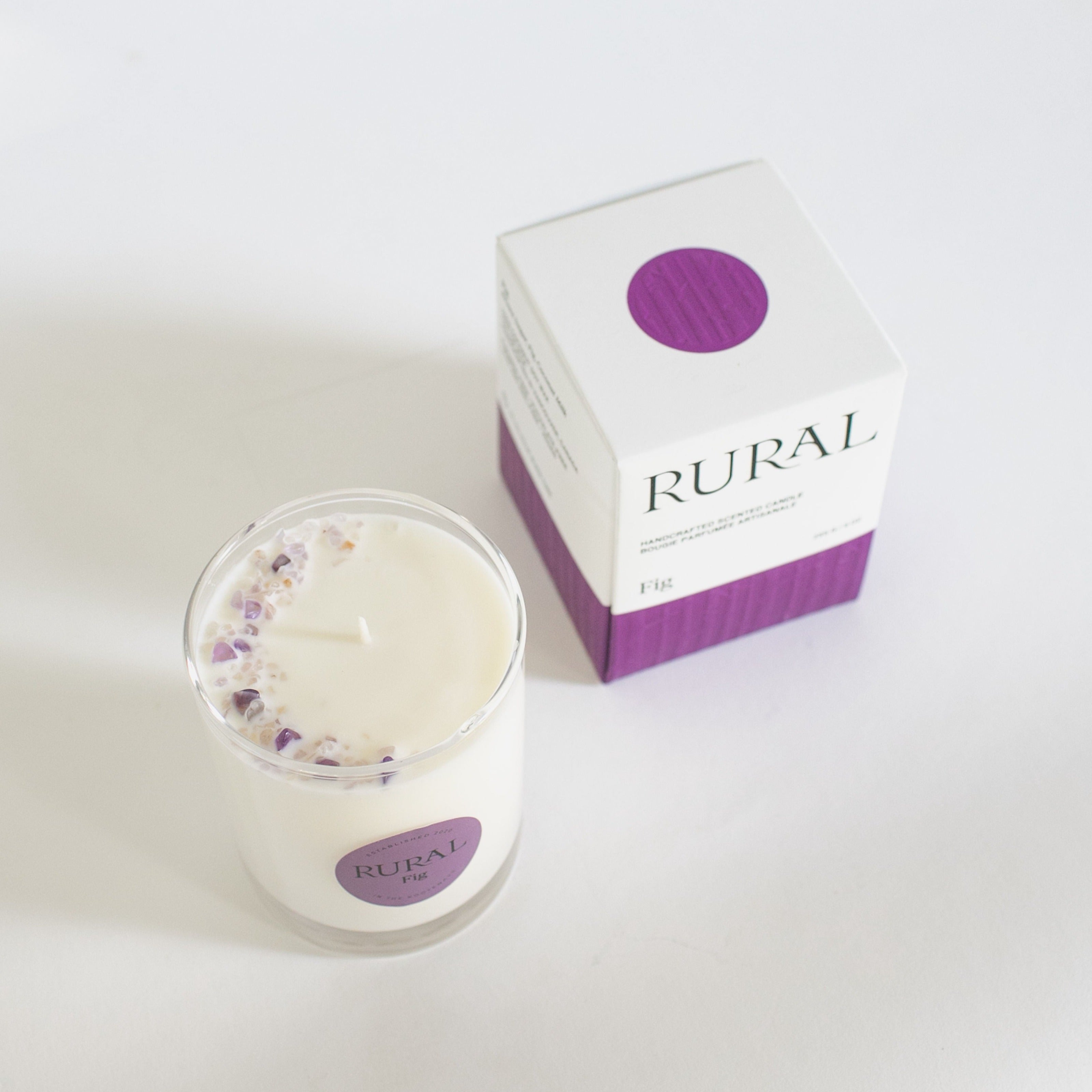 Top view photo of a white candle in a clear glass with a round purple label indicating RURAL Candles Fig Scent beside its matching purple and white elegantly embossed candle gift box. We can see the top of the candle is decorated with small purple amethyst gemstones, artfully arranged in a crescent moon shape.