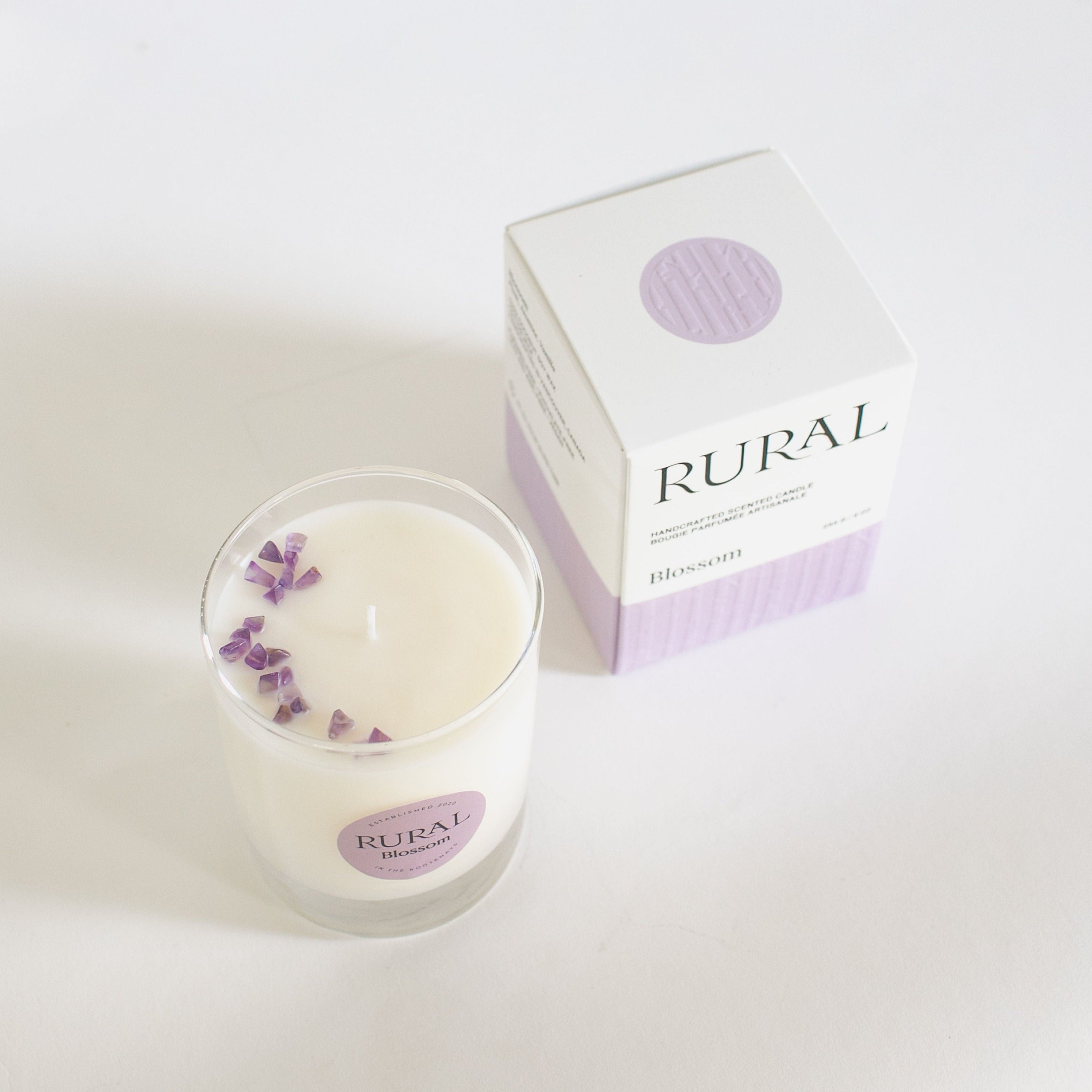 Top view photo of a white candle in a clear glass container with a light purple round RURAL Candles label on it, indicating the scent is called Blossom. The candle top is embellished with a sprinkling of purple amethyst chips, and the candle is sitting beside its matching purple and white, embossed RURAL Candles gift box.