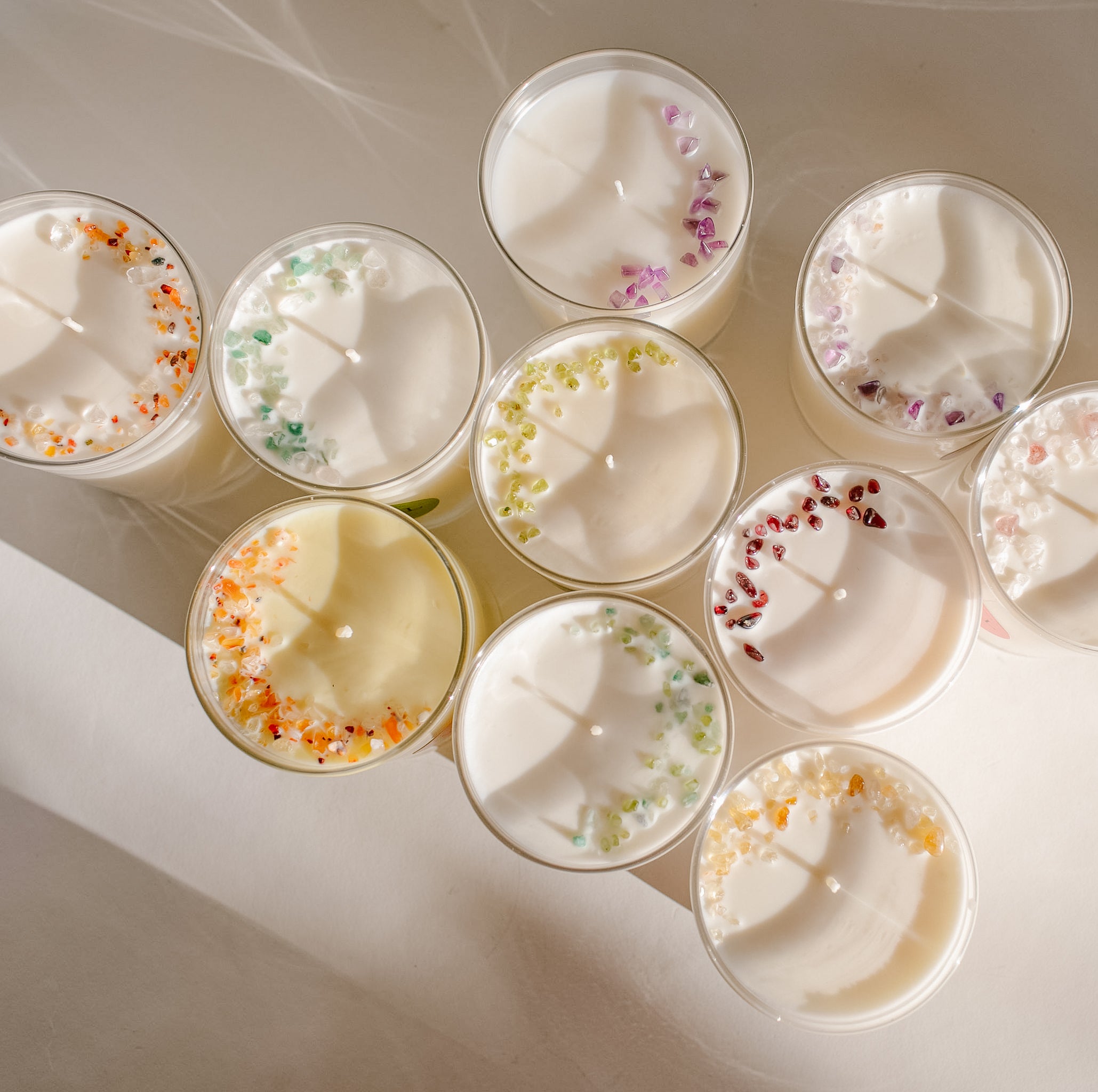 Photo of the top view of a grouping of 11 candles, highlighting the creamy white texture of our coconut-soy wax and the colourful tiny gemstone embellishments, tastefully adorning each candle in a half-moon shape.