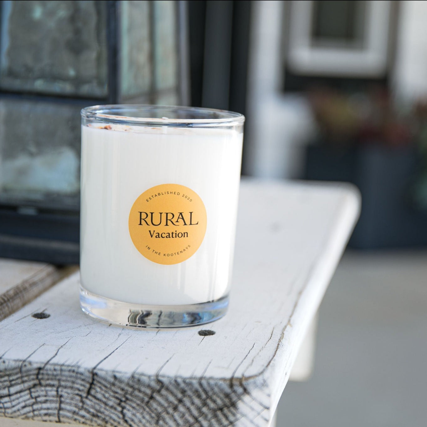 Photo of a white candle in a clear glass container with the round RURAL Candles label on it, indicating the Vacation scent. The candle is sitting on a white rustic wooden bench and the scene is a very calm and inviting.