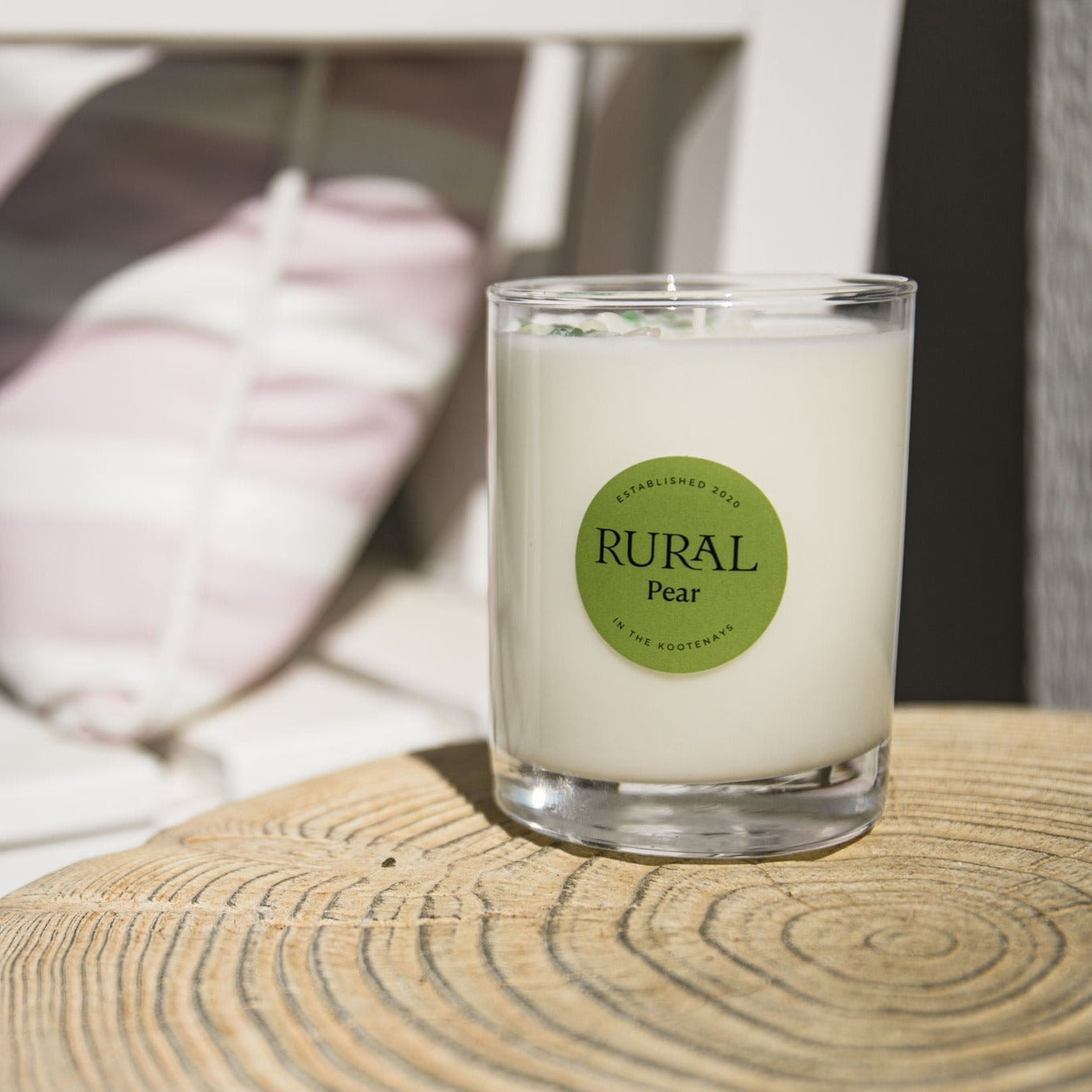 Photo of a white candle in a clear glass with a round green label indicating RURAL Candles Pear Scent. The candle is sitting on a wooden outdoor table on a fresh summer day.
