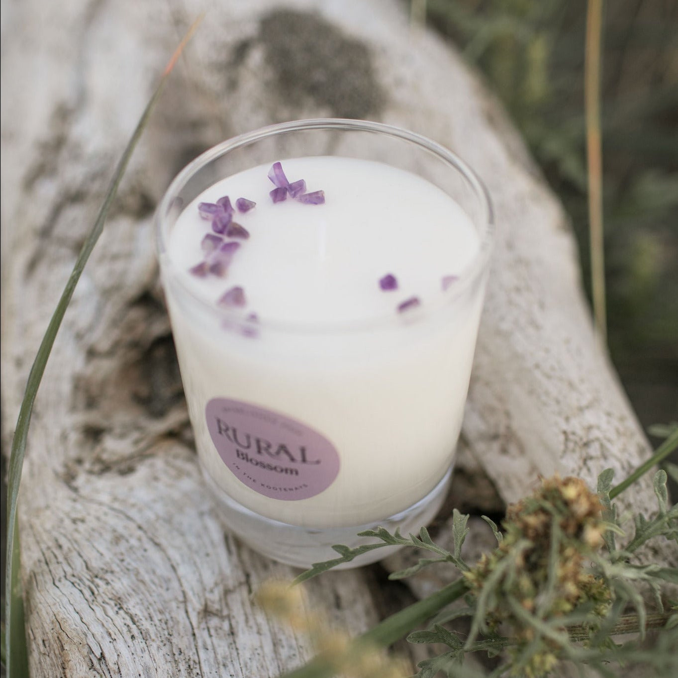 Photo of a white candle in a clear glass container with a light purple round RURAL Candles label on it, indicating the scent is called Blossom. The candle top is embellished with a sprinkling of purple amethyst chips, and the candle is sitting atop a log on the beach.