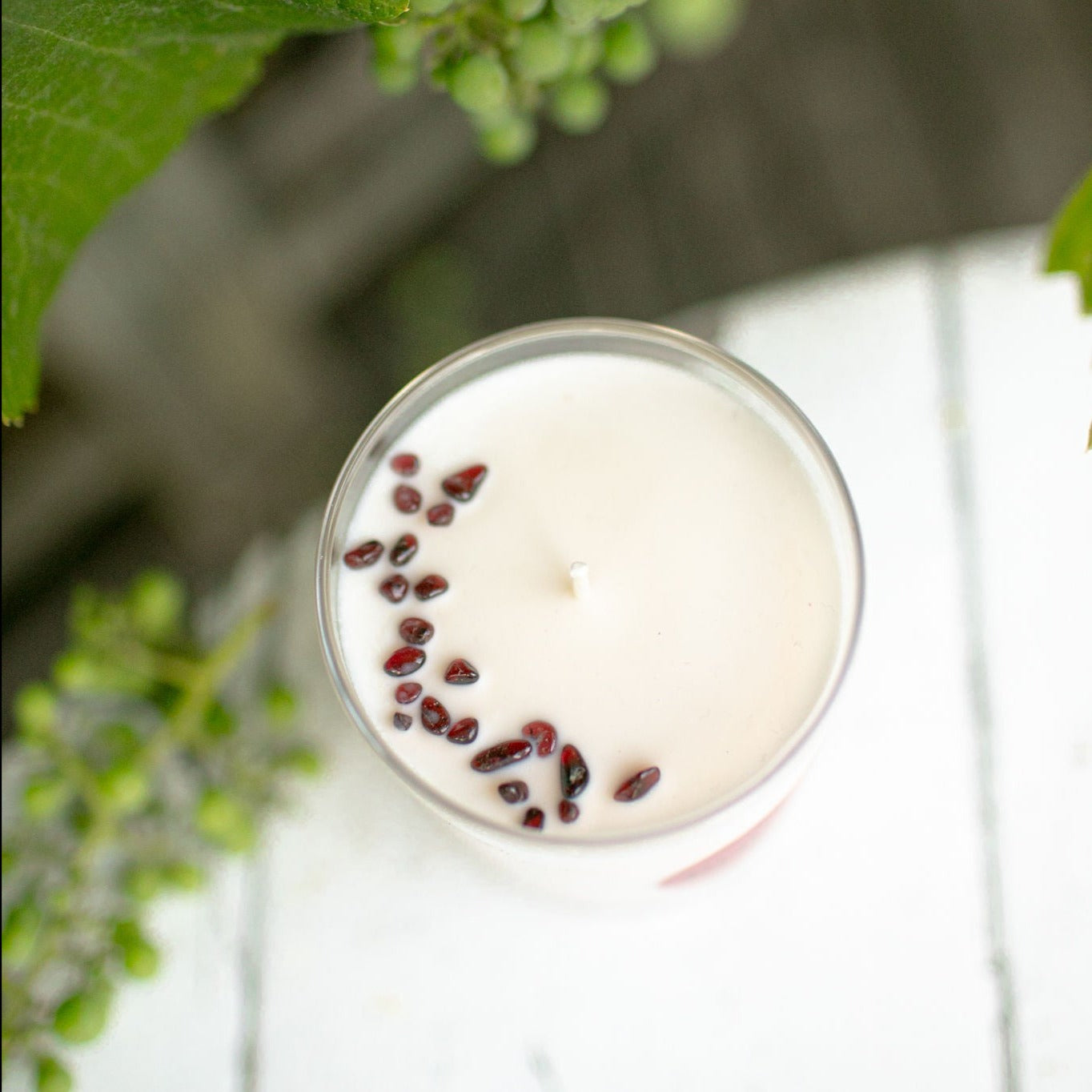 Top view photo of a white candle in a clear glass. This is the RURAL Candles Pomegranate candle with red garnet gemstones. The candle is on a white table surrounded by green grapes, looking like a fresh summer day in the garden.