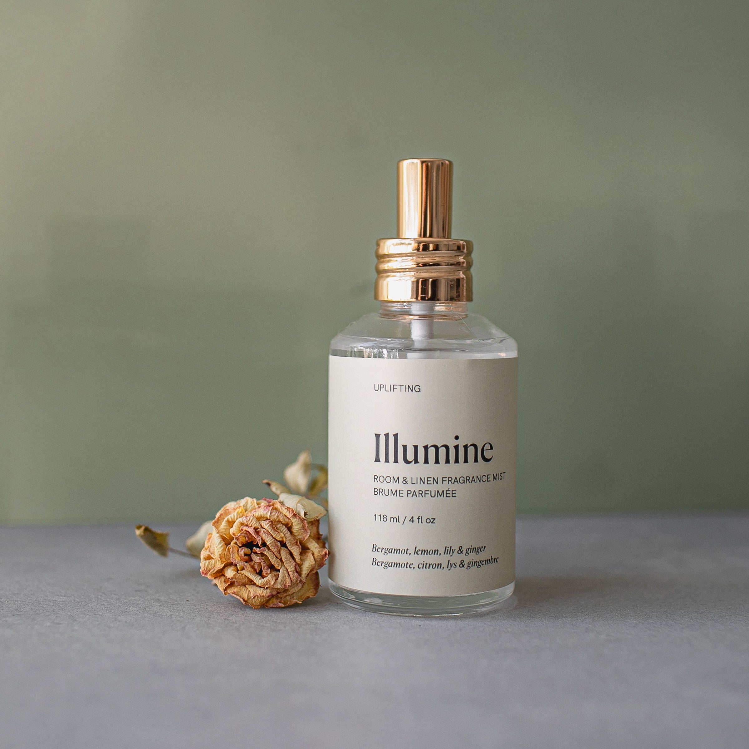 Bottle of 'Illumine' room & linen fragrance mist with a gold cap on a gray surface against a green background.