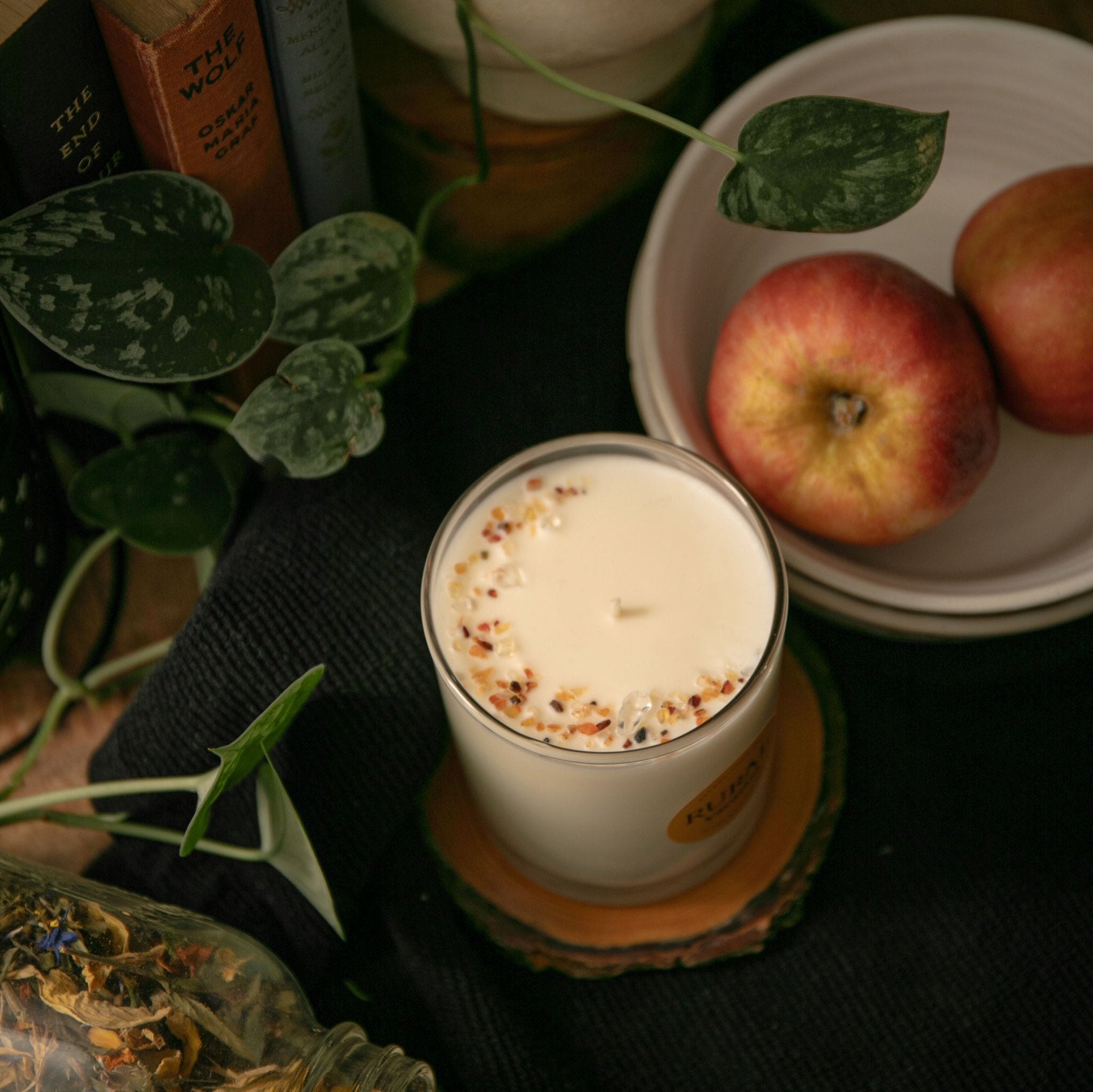 Top view photo of a coconut soy candle in a clear glass container with the round RURAL Candles label on it, indicating the Vacation scent. The top of the candle is sprinkled with carnelian gemstones and clear quartz crystals, arranged in a crescent moon shape, and the scene is a cozy and mystical kitchen vibe, with a bowl of red apples, jar of dried flowers, green plants and magical books in the background.