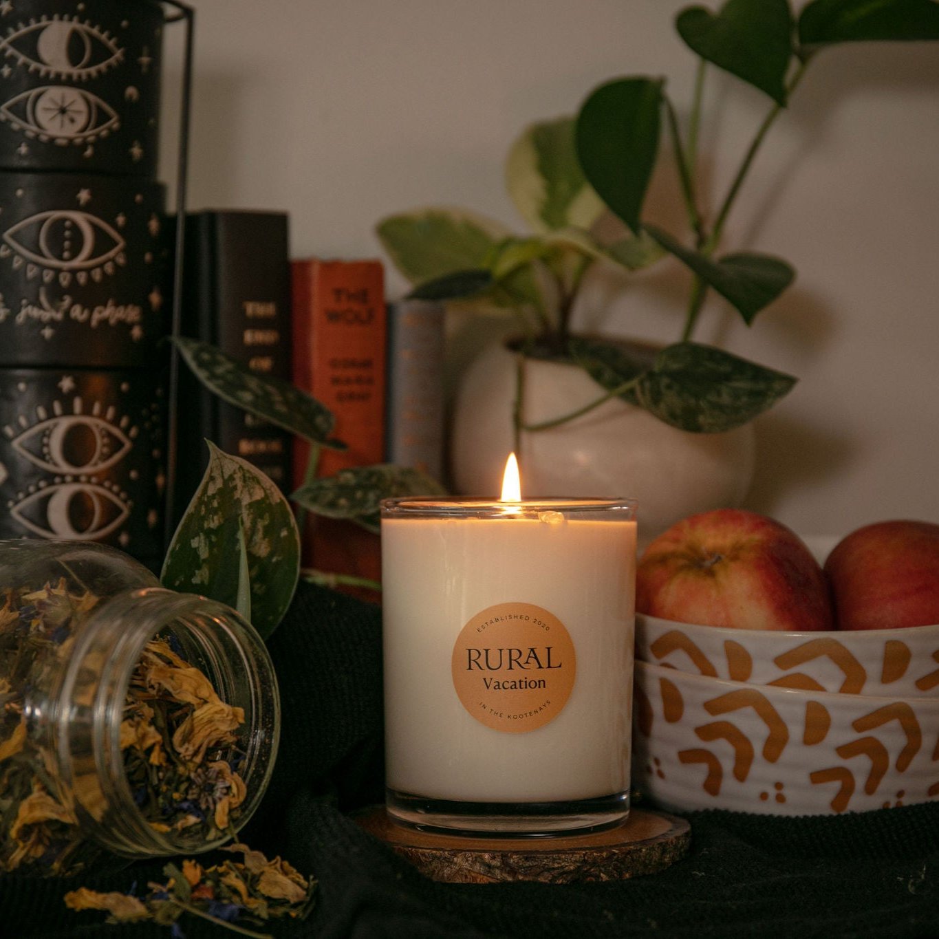 Photo of a coconut soy candle in a clear glass container with an orange circle RURAL Candles label on it, indicating the Vacation scent. The candle is lit and the scene is a cozy and mystical kitchen vibe, with a bowl of red apples, jar of dried flowers, green plants and magical books in the background.