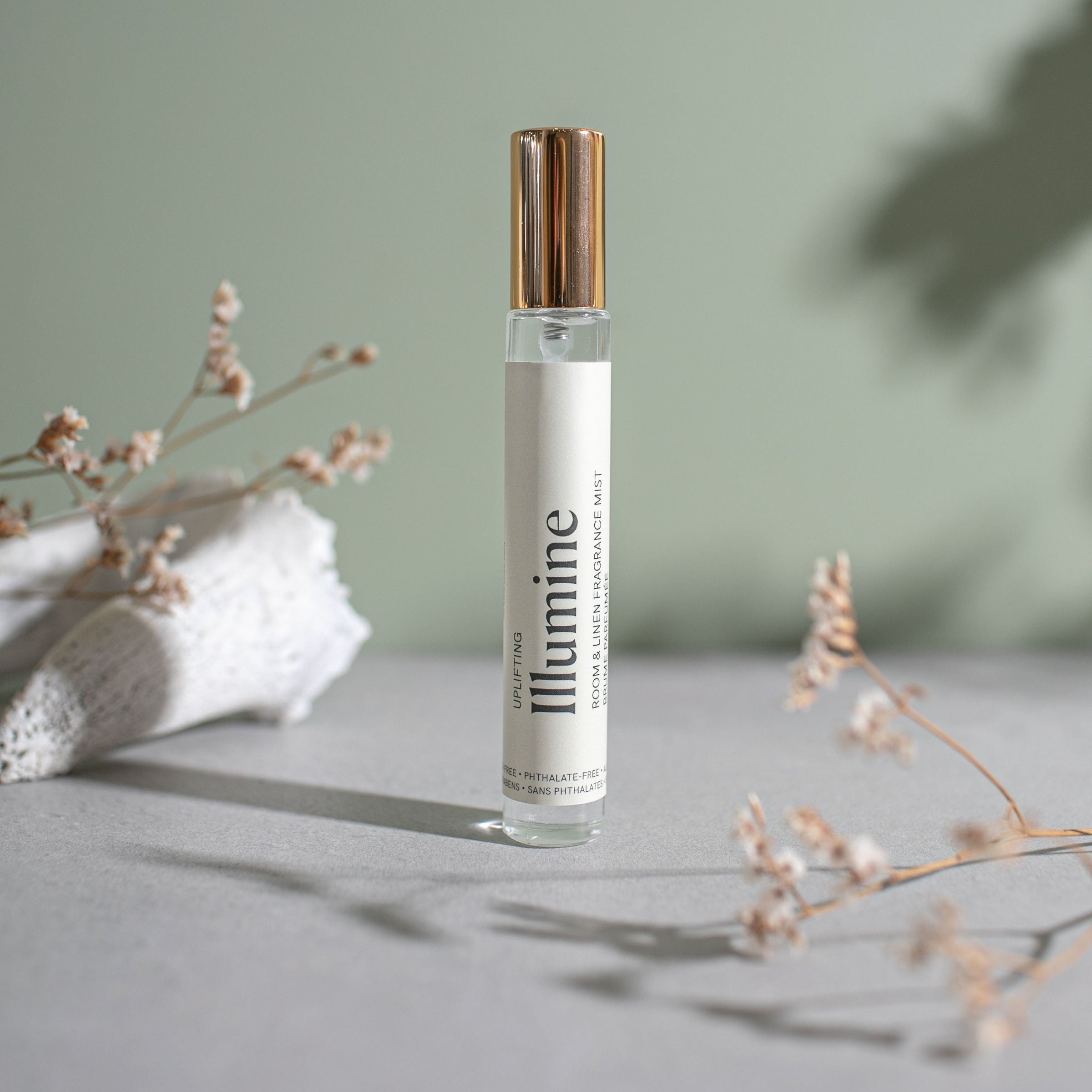 Small spray bottle with a gold cap labelled 'Illumine' on a neutral background with dried flowers.