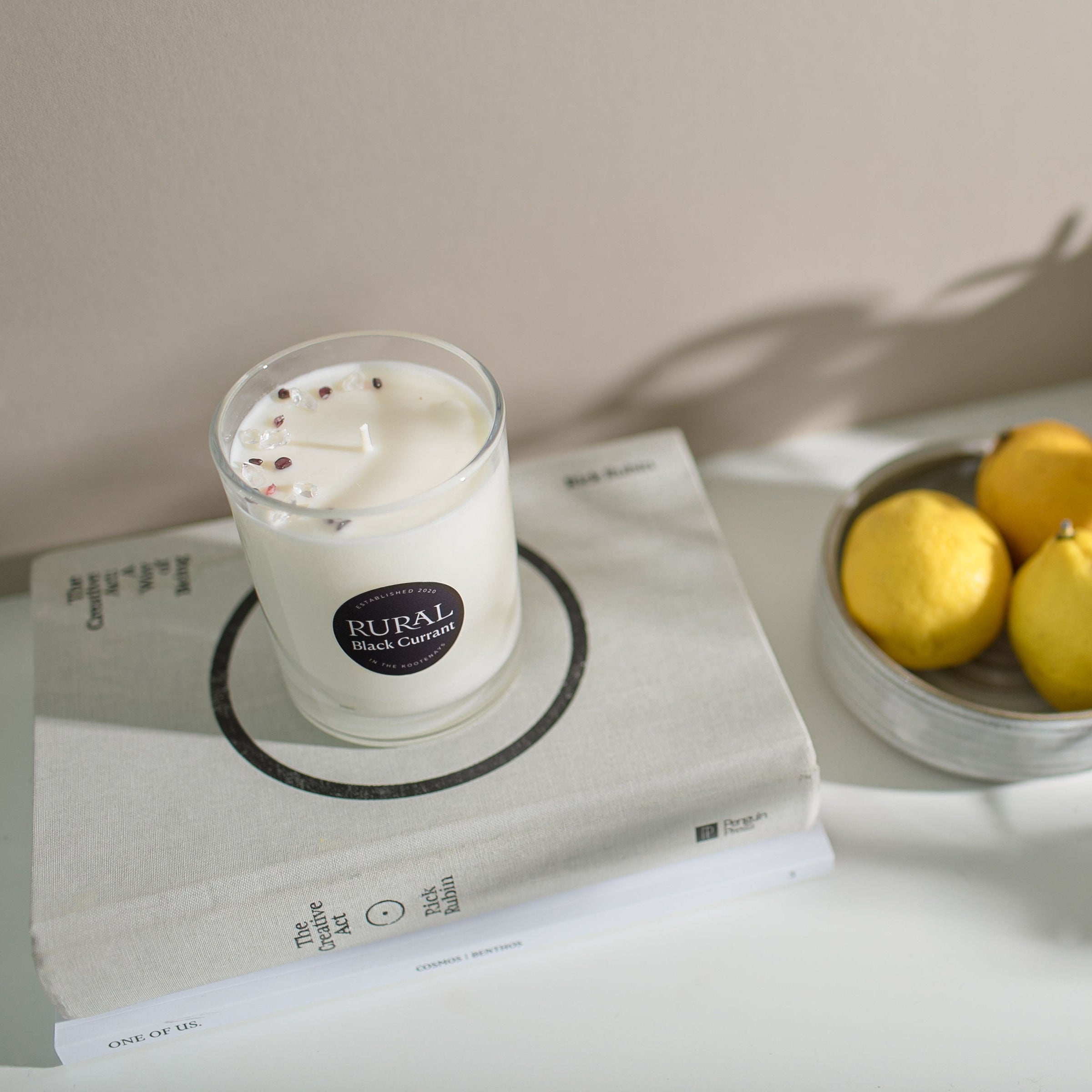 White candle on a book next to a bowl of lemons and a plant.