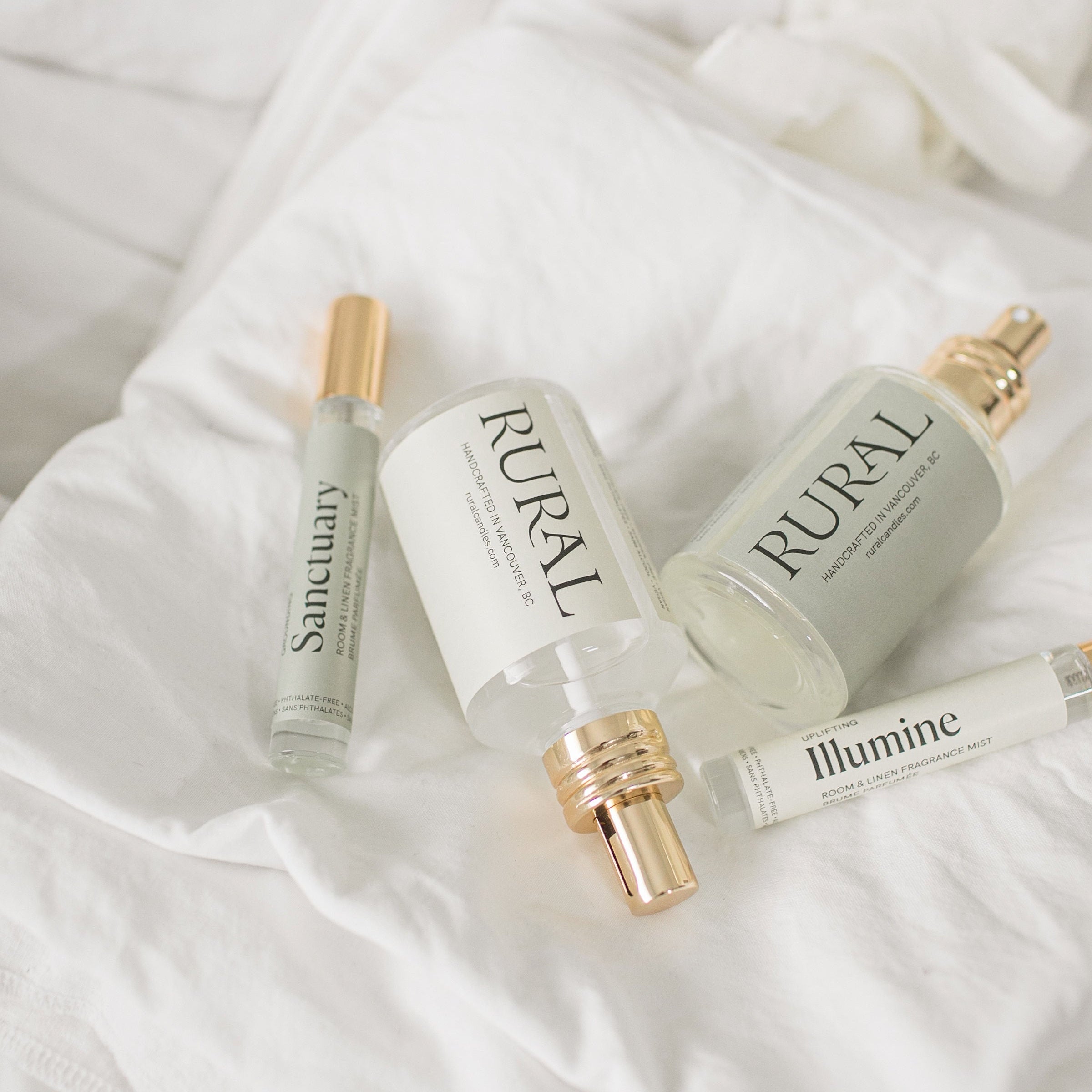 Large and small glass bottles labelled 'Rural', 'Sanctuary', and 'Illumine' on a white fabric background