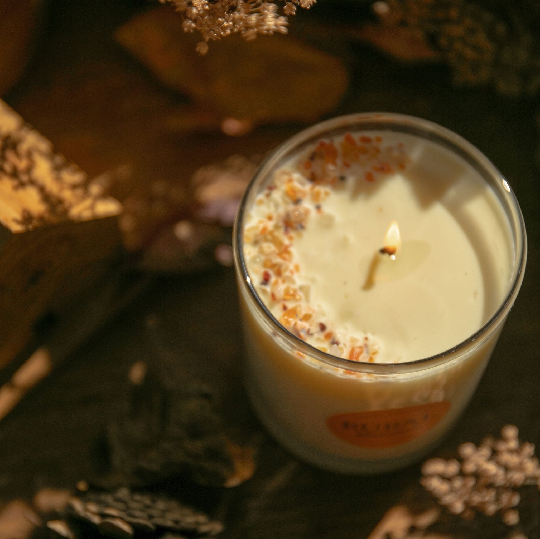 Top view photo of a RURAL Candles Mandarin scented candle in a clear glass with a round orange label. We can see the top of the candle is decorated with small orange carnelian gemstones and clear quartz crystals, artfully arranged in a crescent moon shape. The candle has a gentle flame and is on a wooden table with pinecones and dried flower decor for a cozy Fall feel.