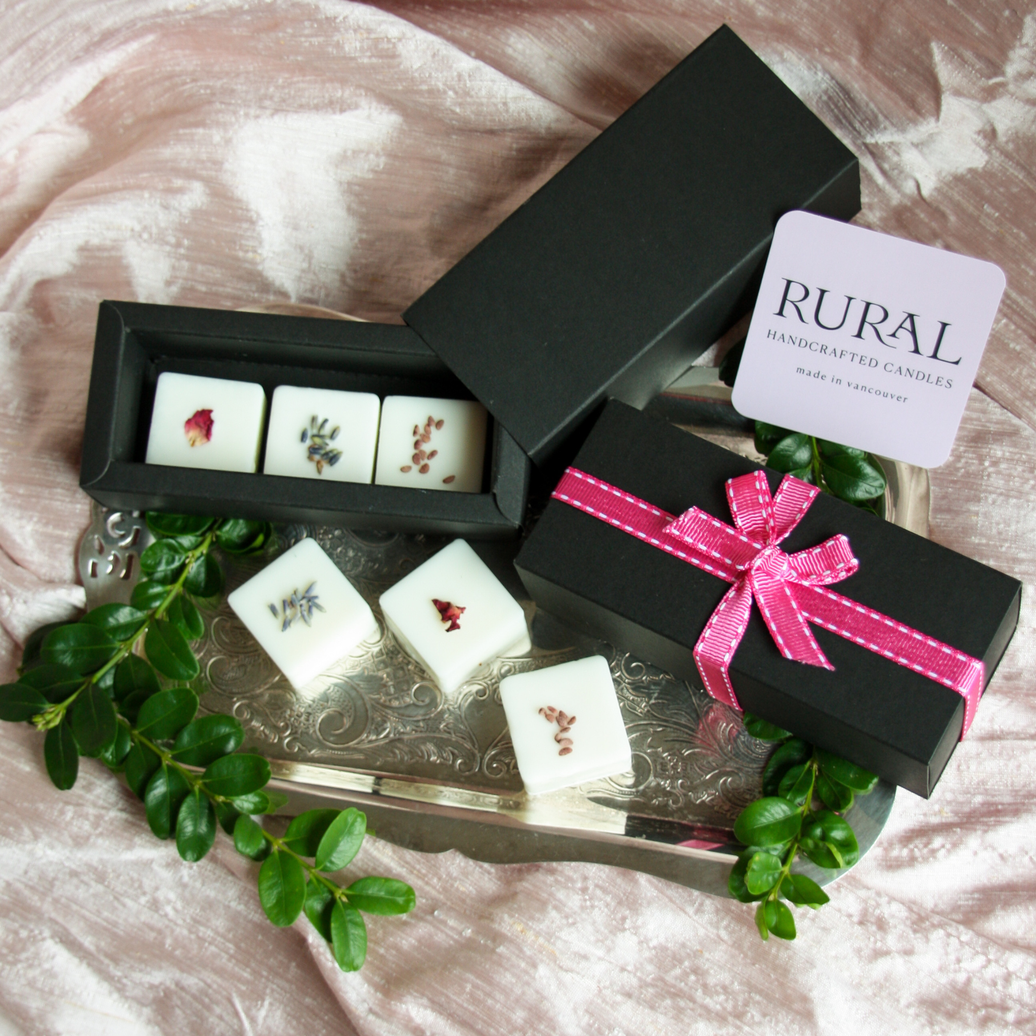 Photo of a black gift box with a pink ribbon opened to show six square coconut soy wax melts handcrafted by RURAL Candles, decorated with red and purple flower petals and tiny blackberry seeds. All artfully displayed on a silver platter, pink silk fabric and sprigs of green in the background.