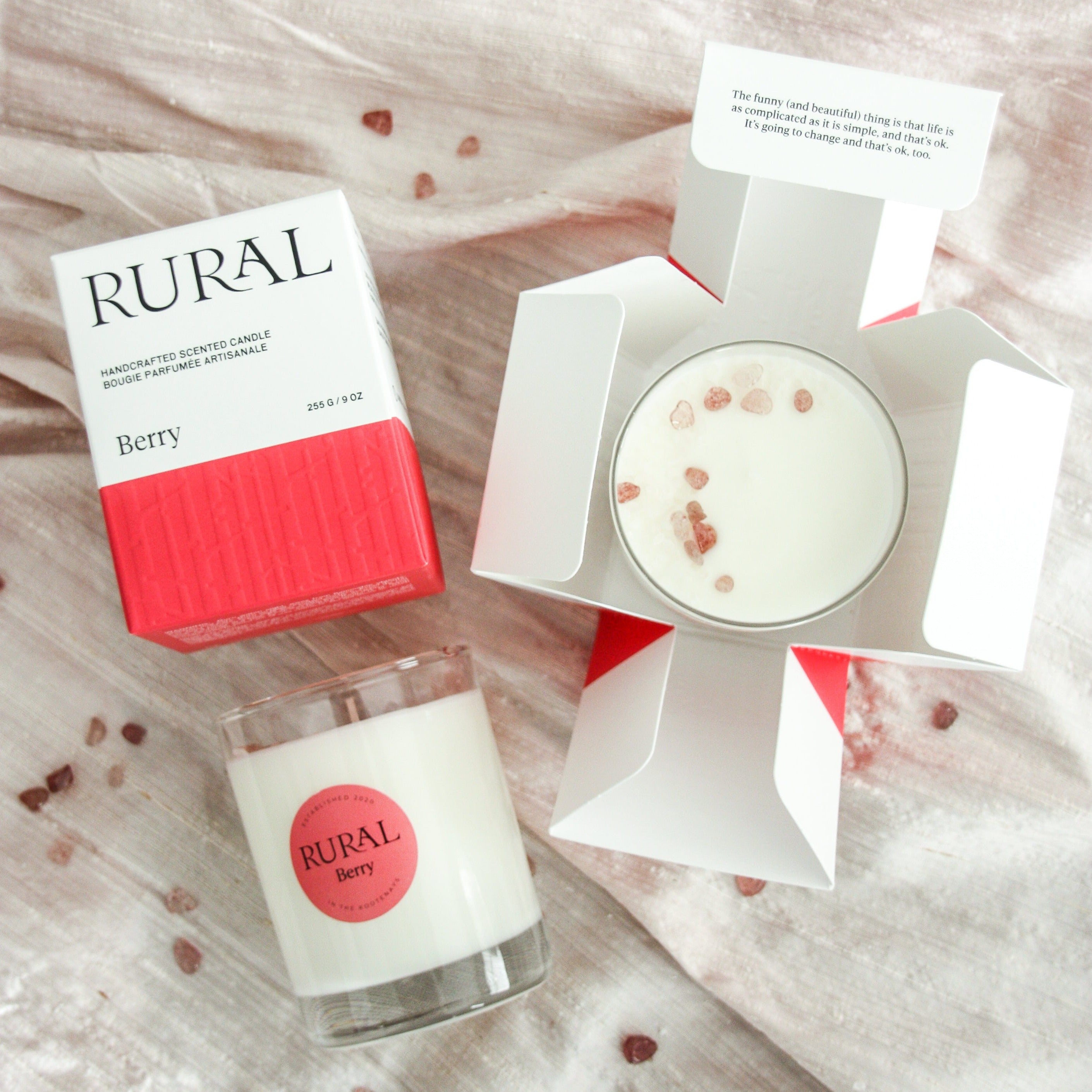 Photo of two white candles in clear glass containers with a pink round RURAL Candles label on, indicating its Berry scent. The candles are on a pink silk fabric. The tops of the candles are sprinkled with various shades of pink strawberry quartz crystals. Alongside is a matching pink and white Berry Candle gift box with the RURAL Candles logo on it. The gift box opens like a flower.