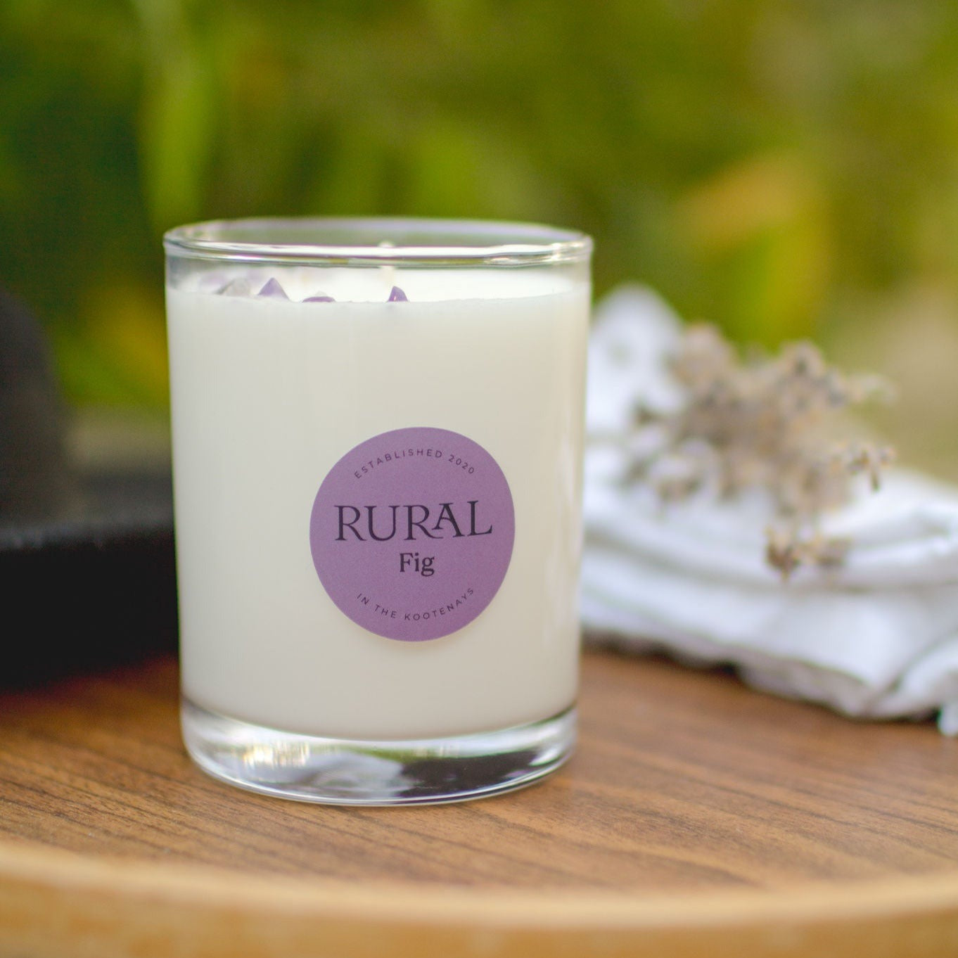 Photo of a white candle in a clear glass with a round purple label indicating RURAL Candles Fig scent. The candle is sitting on a wooden table in a calming garden setting.