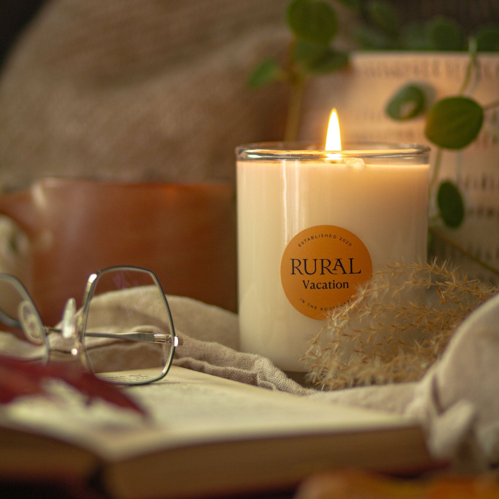 Photo of a coconut soy candle in a clear glass container with the round RURAL Candles label on it, indicating the Vacation scent. The candle is lit and the scene is a cozy and reading nook vibe, with a pair of metal rimmed eyeglasses, an open book, green plants and dried flowers in the background.