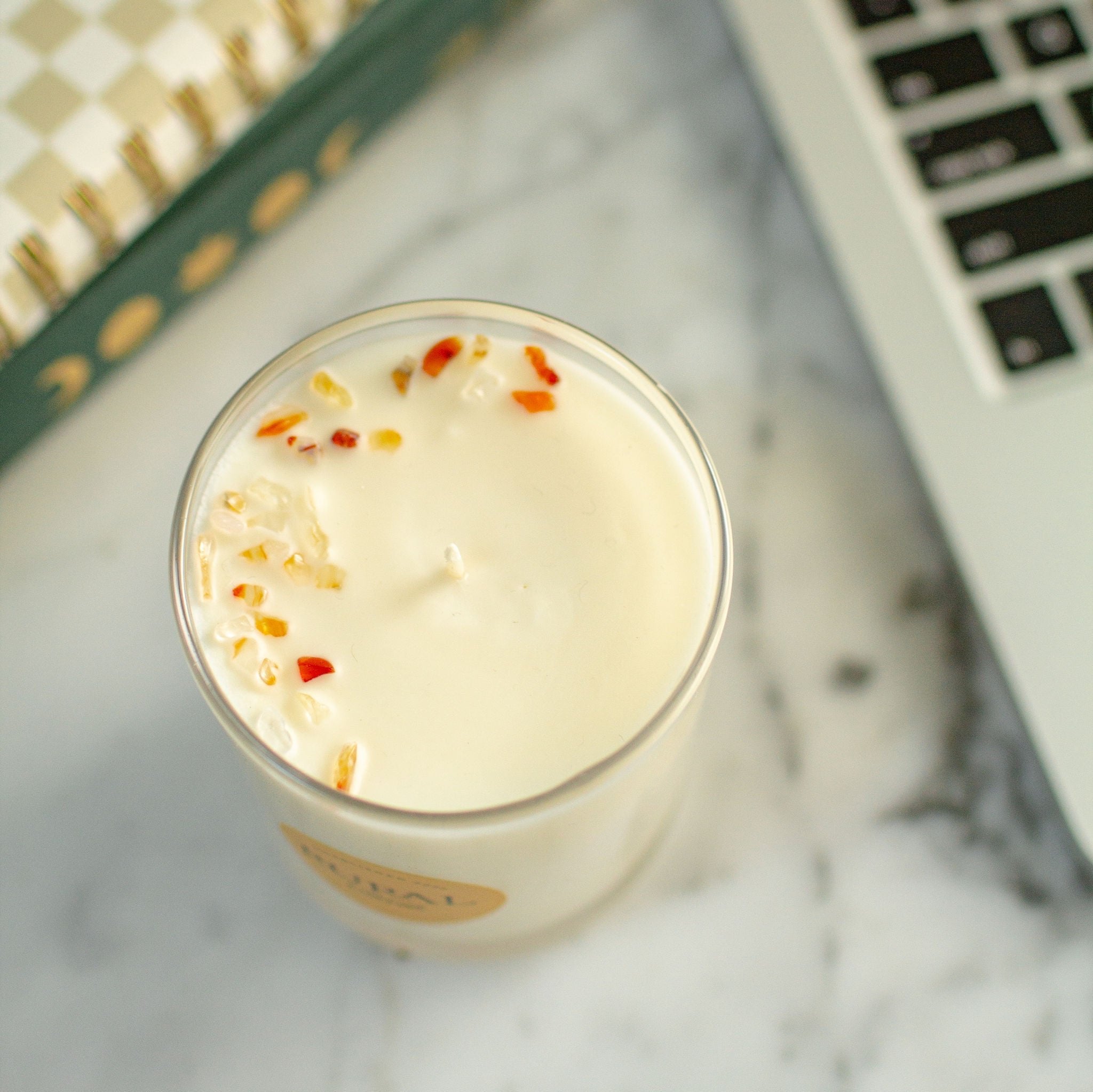 Top view photo of a white candle in a clear glass container with a yellow round RURAL Candles label on it. We can see the top of the candle is embellished with various shades of orange carnelian gemstones, artfully arranged in a half-moon shape.