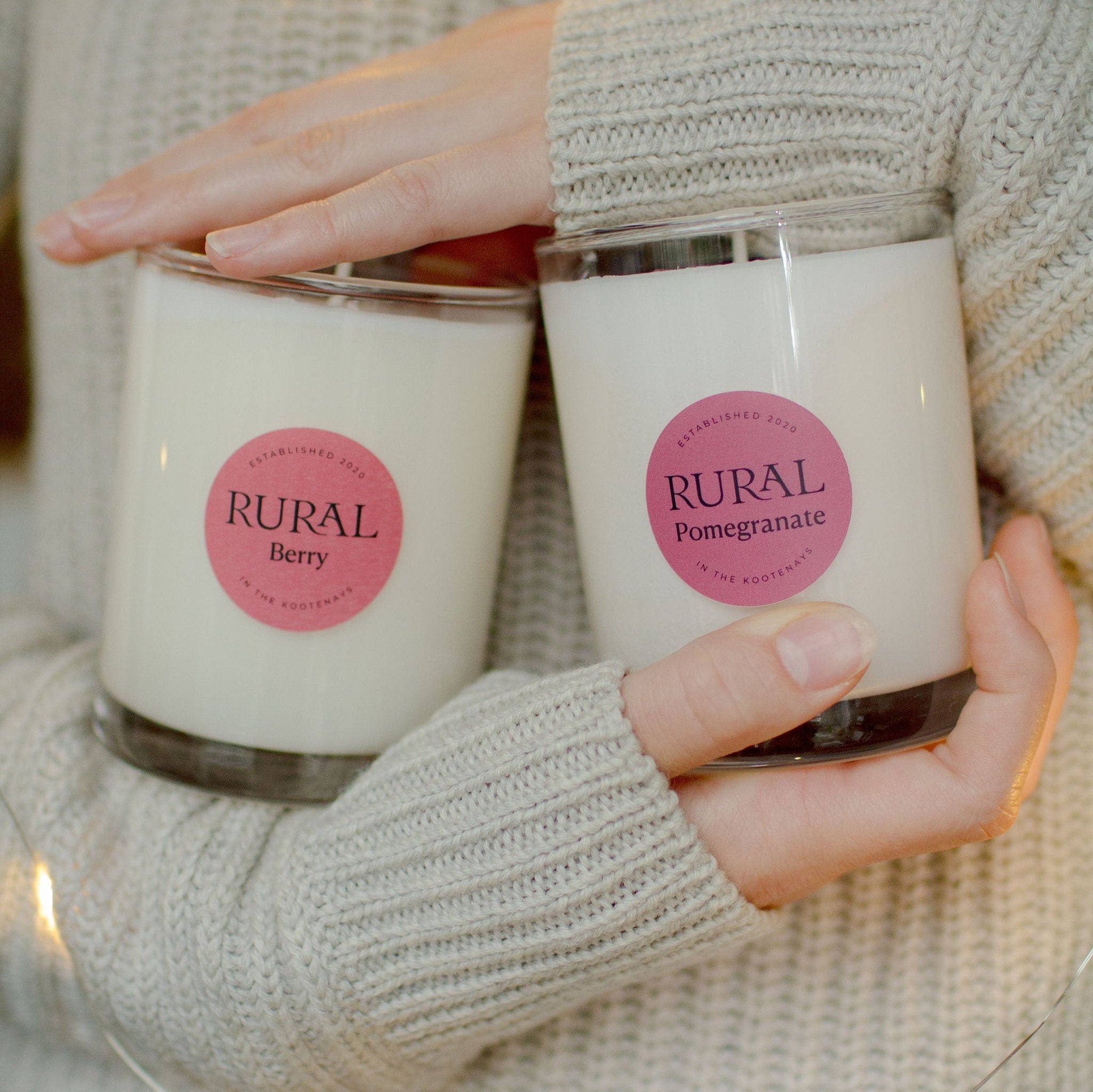 Two white candles in clear glass containers with pink and red round labels, indicating RURAL scented candles in Berry and Pomegranate fragrances. The candles are being cradled in a girls arms, she's wearing a cozy knit sweater, giving winter comfort vibes.