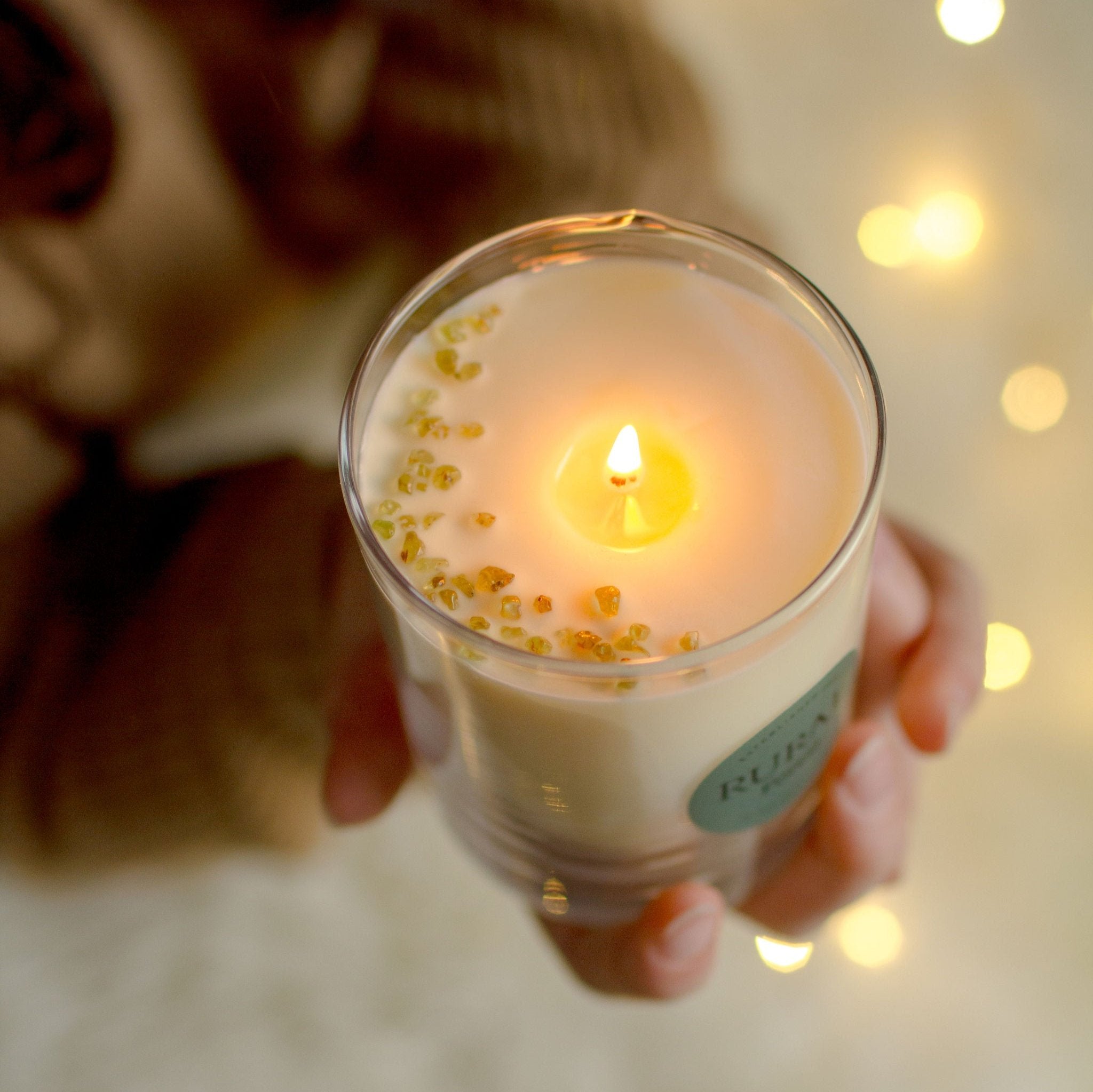 Top view photo of a white candle in a clear glass with a round green label indicating RURAL Candles Forest Scent. The candle is lit and held by a girl's hands, she's wearing a cozy sweater and fairy lights are twinkling in the background, giving a magical cozy vibe. We can see the top of the candle is decorated with small green olivine crystals, arranged in a crescent moon shape.