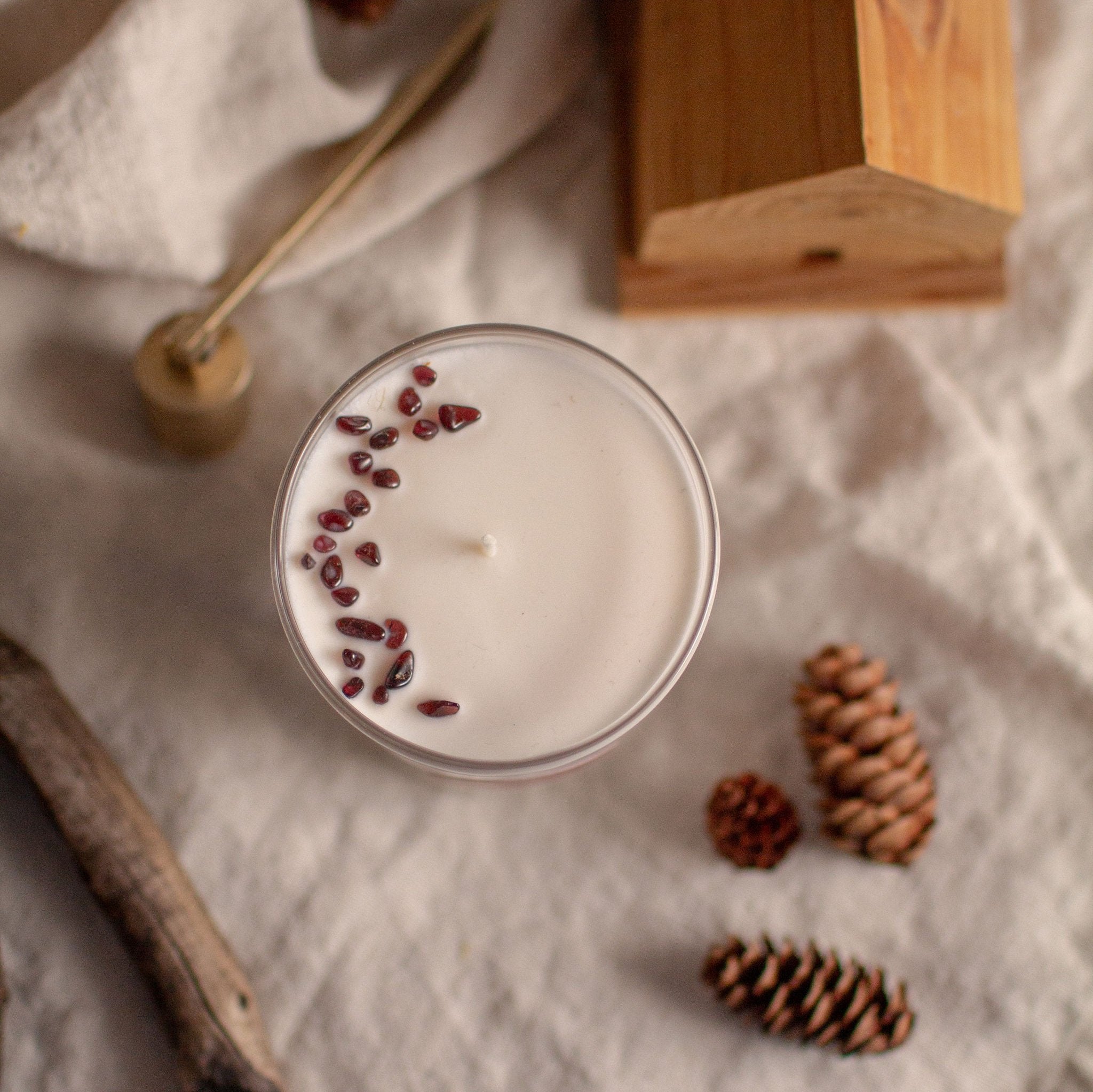 Top view photo of a white candle in a clear glass. This is the Pomegranate candle with gemstones. The candle is on a linen tablecloth with pinecones and wood decor, creating a soothing, cozy evening vibe.