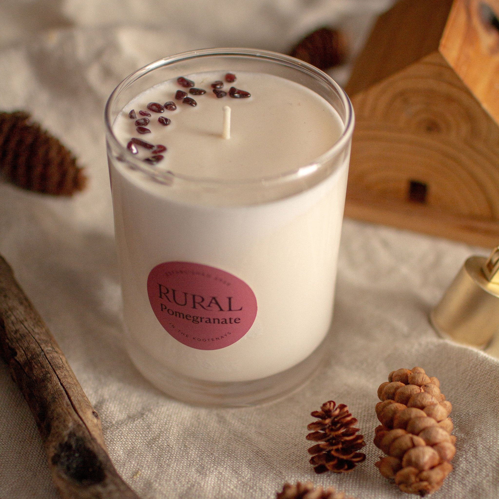 Photo of a white candle in a clear glass with a round red label indicating RURAL Candles Pomegranate Scent. We can see the top of the candle is decorated with small red garnet gemstones, arranged in a crescent moon shape. The candle is on a linen tablecloth with pinecones and fairy lights creating a cozy space.