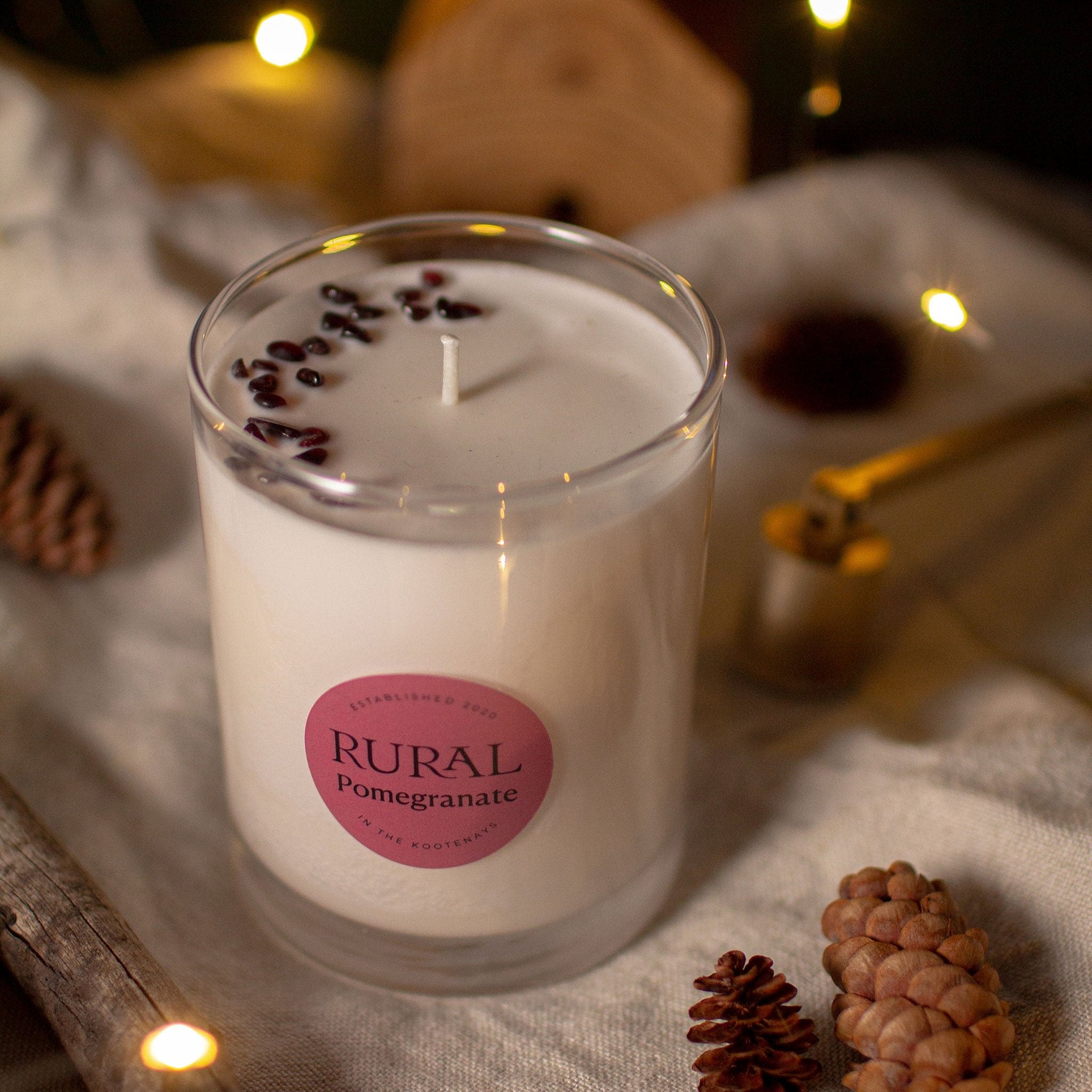 Photo of a white candle in a clear glass with a round red label indicating RURAL Candles Pomegranate Scent. We can see the top of the candle is decorated with small red garnet gemstones, arranged in a crescent moon shape. The candle is on a linen tablecloth with pinecones and fairy lights creating a cozy space.