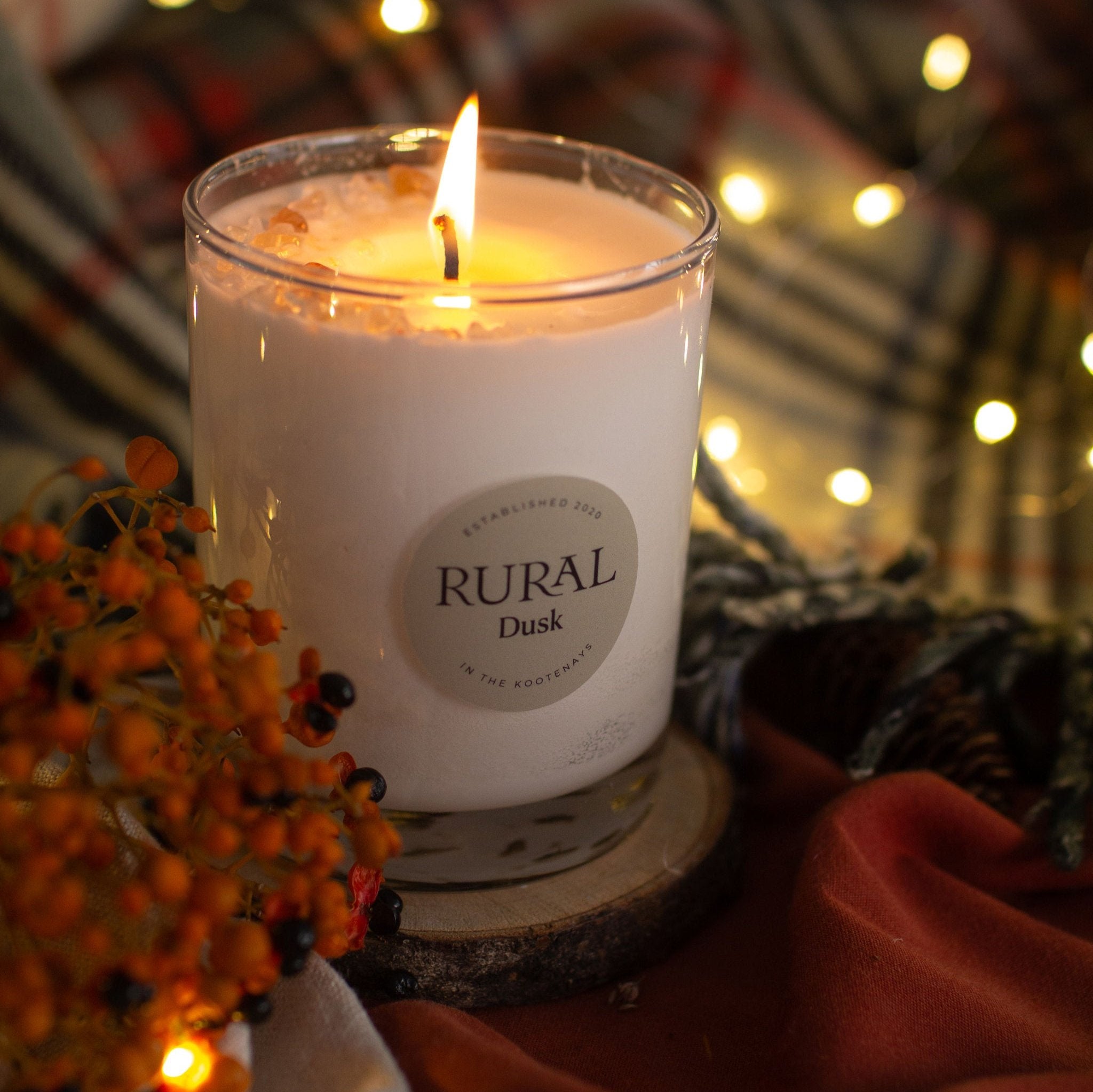 Photo of a Dusk scented RURAL Candle in a clear glass. The candle is lit and sitting on a wooden coaster and tartan blankets with orange berries, woollen fabrics and fairy lights surrounding it. The scene is very cozy and warm feeling for a Fall or winter evening.