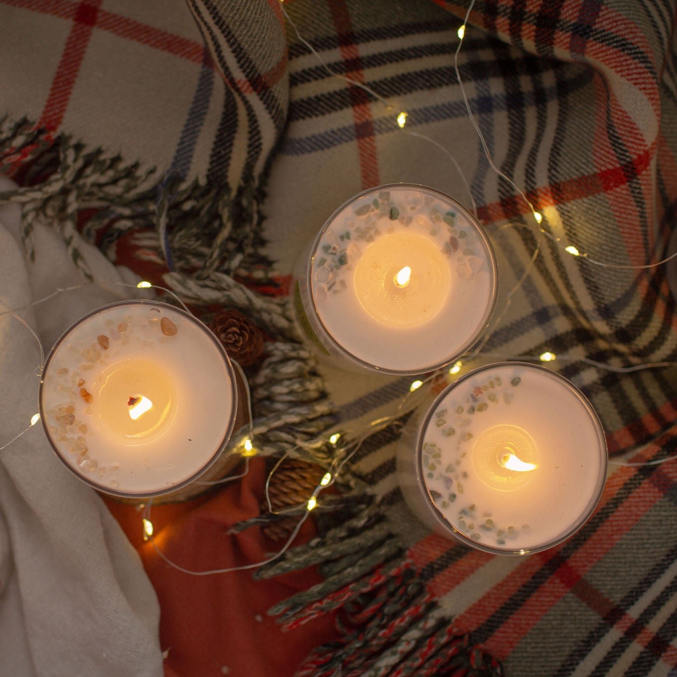 Tiop view of a group of three candles in clear glass with colorful gemstones on top in a crescent moon pattern. The candles are on an orange and black tartan blanket with fairy lights weaving through, creating a warm, cozy and inviting space.