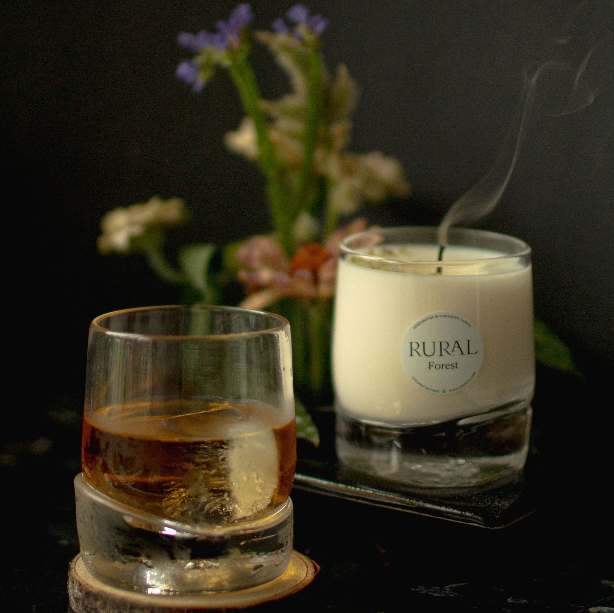 Photo of two very unique hand-blown, heavy-bottom glass whiskey tumblers. One has been handcrafted into a RURAL Candles Forest scented candle, and the other holds a drink, which could be whiskey on the rocks. In the background is an exotic bouquet of flowers and natural wood and black leather decor. The candle appears to be just blown out, with a plume of smoke gently rising above it, providing a sensual, warm and inviting after hours mood.
