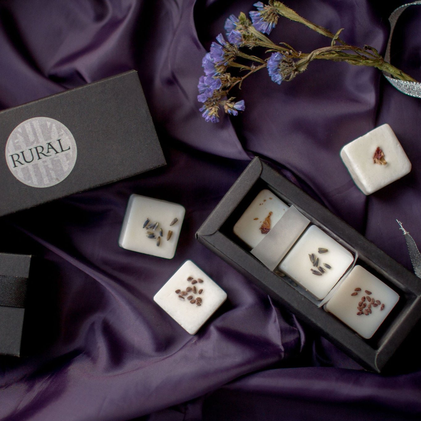 Photo of a black gift box with six square coconut soy wax melts handcrafted by RURAL Candles, decorated with dried flower petals and berry seeds. All artfully displayed on a purple satin cloth with purple and green floral stems alongside.
