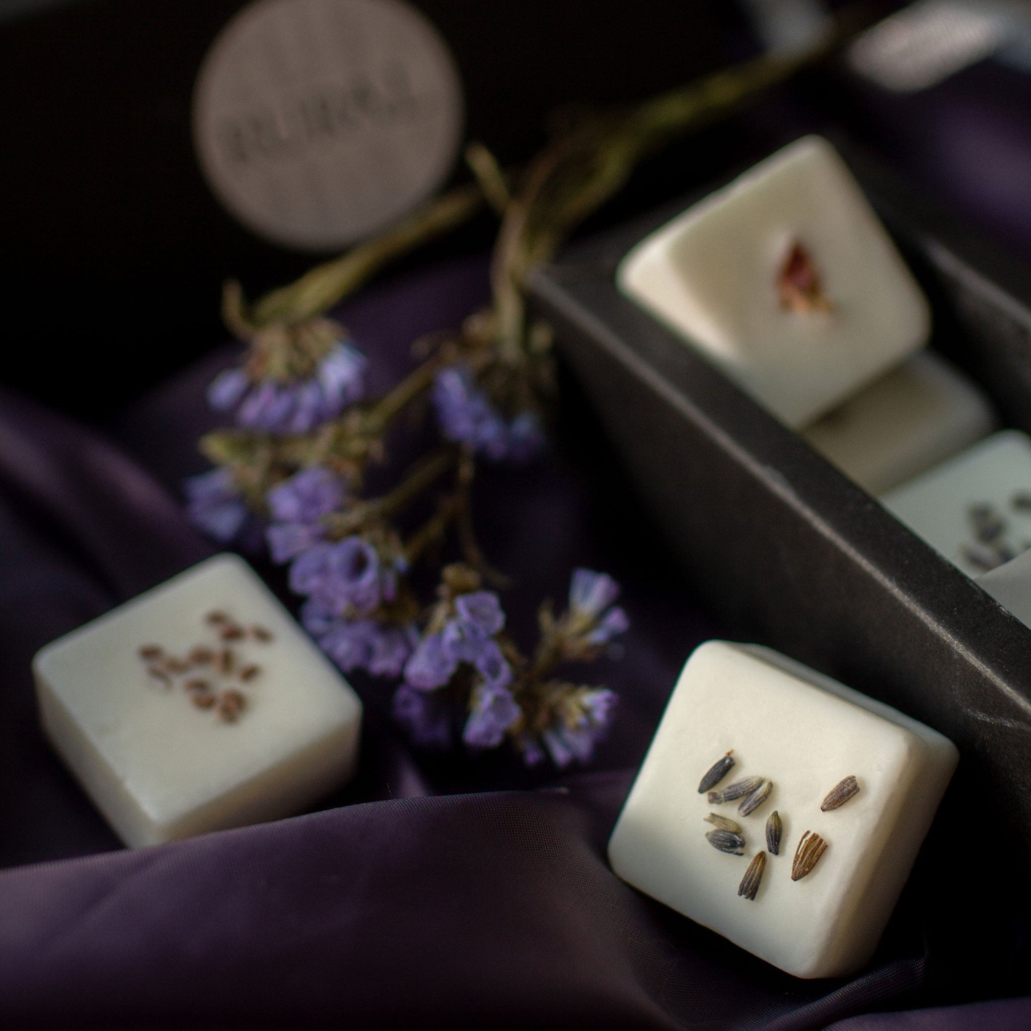 Close-up photo of a black gift box of square coconut soy wax melts handcrafted by RURAL Candles, decorated with dried flower petals and berry seeds. All artfully displayed on a purple satin cloth with purple and green floral stems in the background.