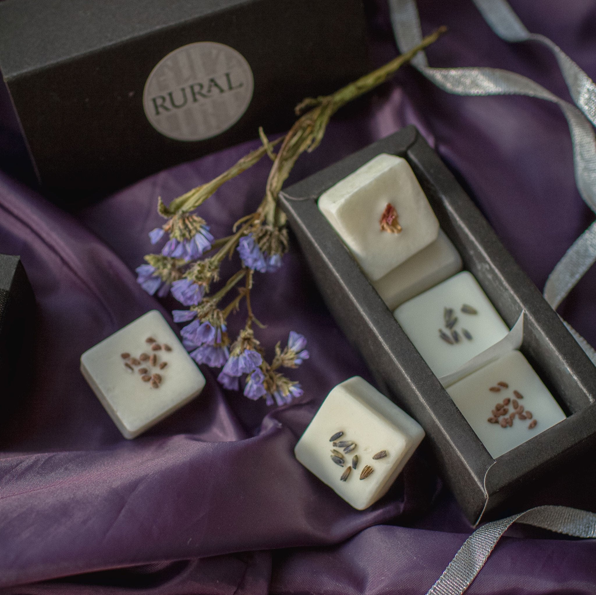 Photo of a black gift box with six square coconut soy wax melts handcrafted by RURAL Candles, decorated with dried flower petals and berry seeds. All artfully displayed on a purple satin cloth with purple and green floral stems alongside.